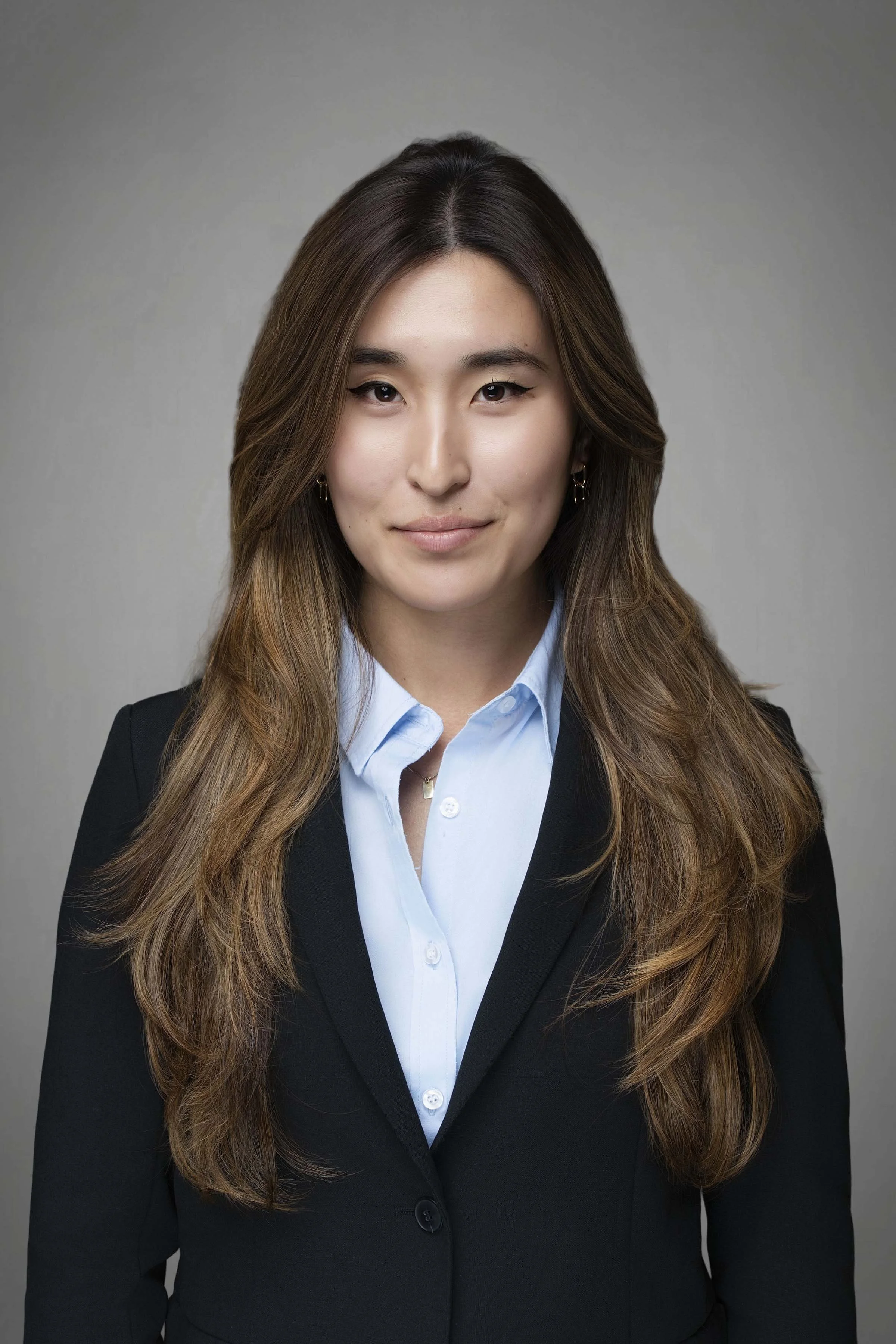 Businesswoman with long brown hair, wearing a black blazer and light blue dress shirt, against a gray background.