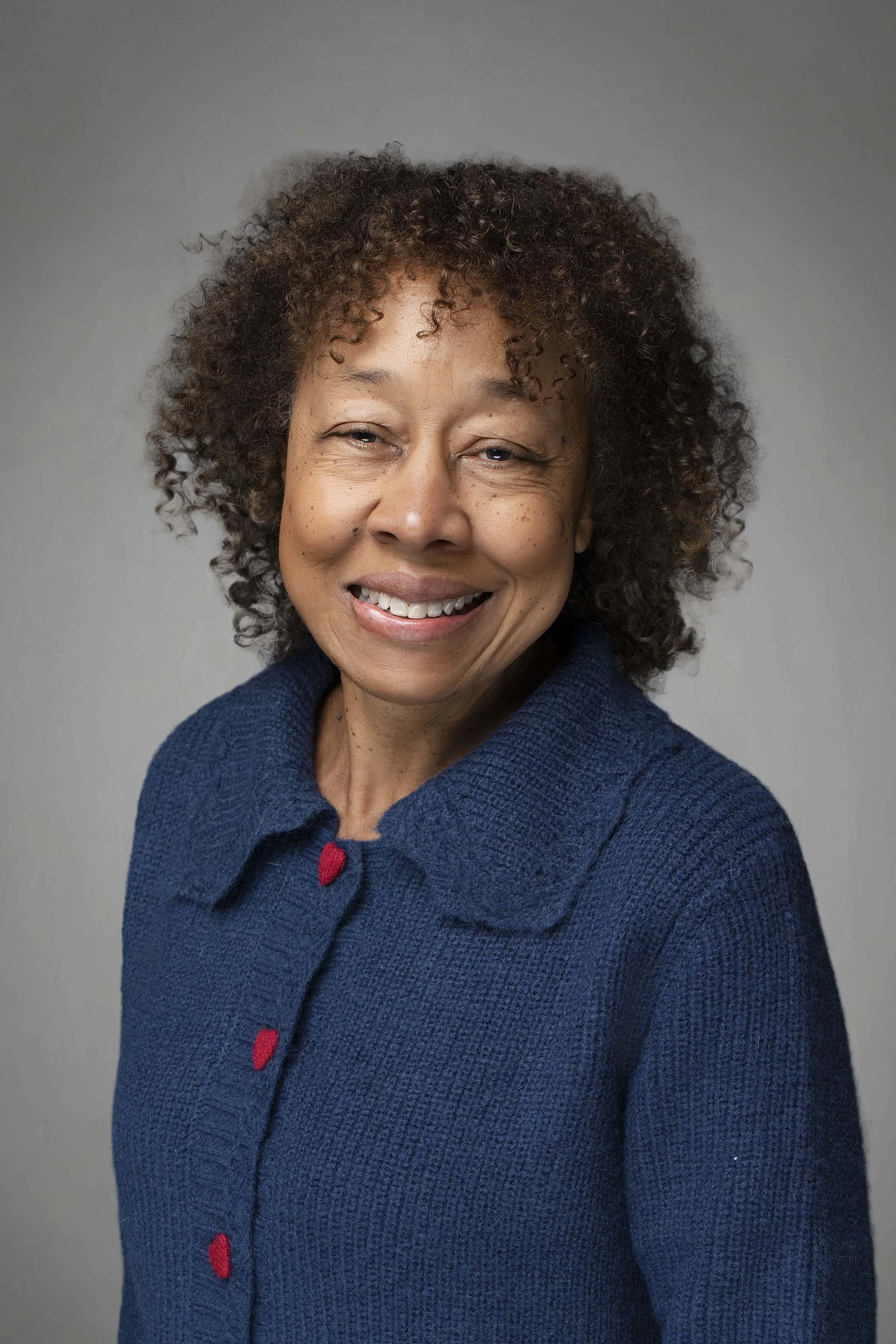 A smiling middle-aged woman with curly hair wearing a blue sweater against a gray background.