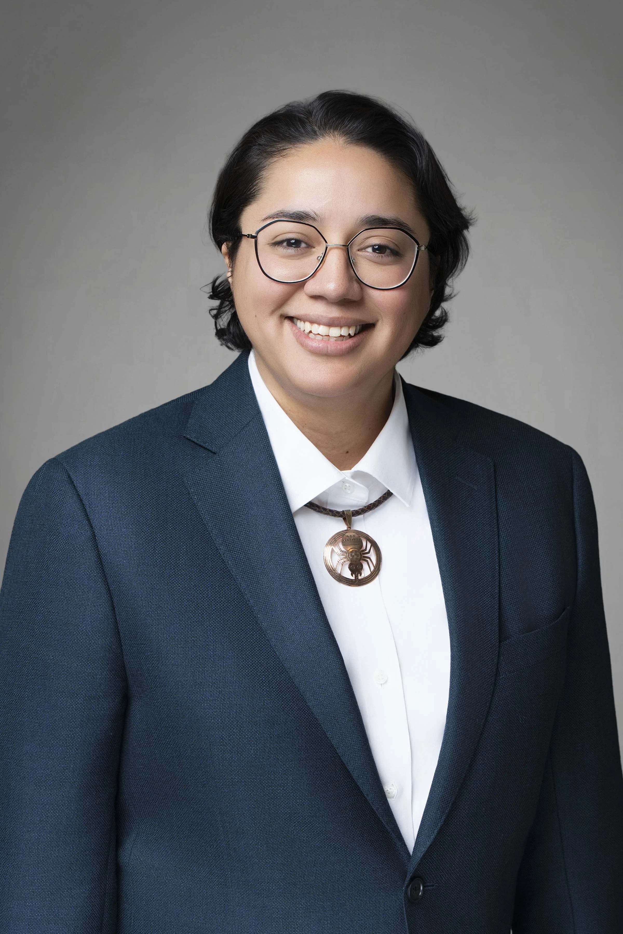 A woman with short black hair, glasses, and a smile, wearing a dark blue blazer, white shirt, and a necklace with a large circular pendant.