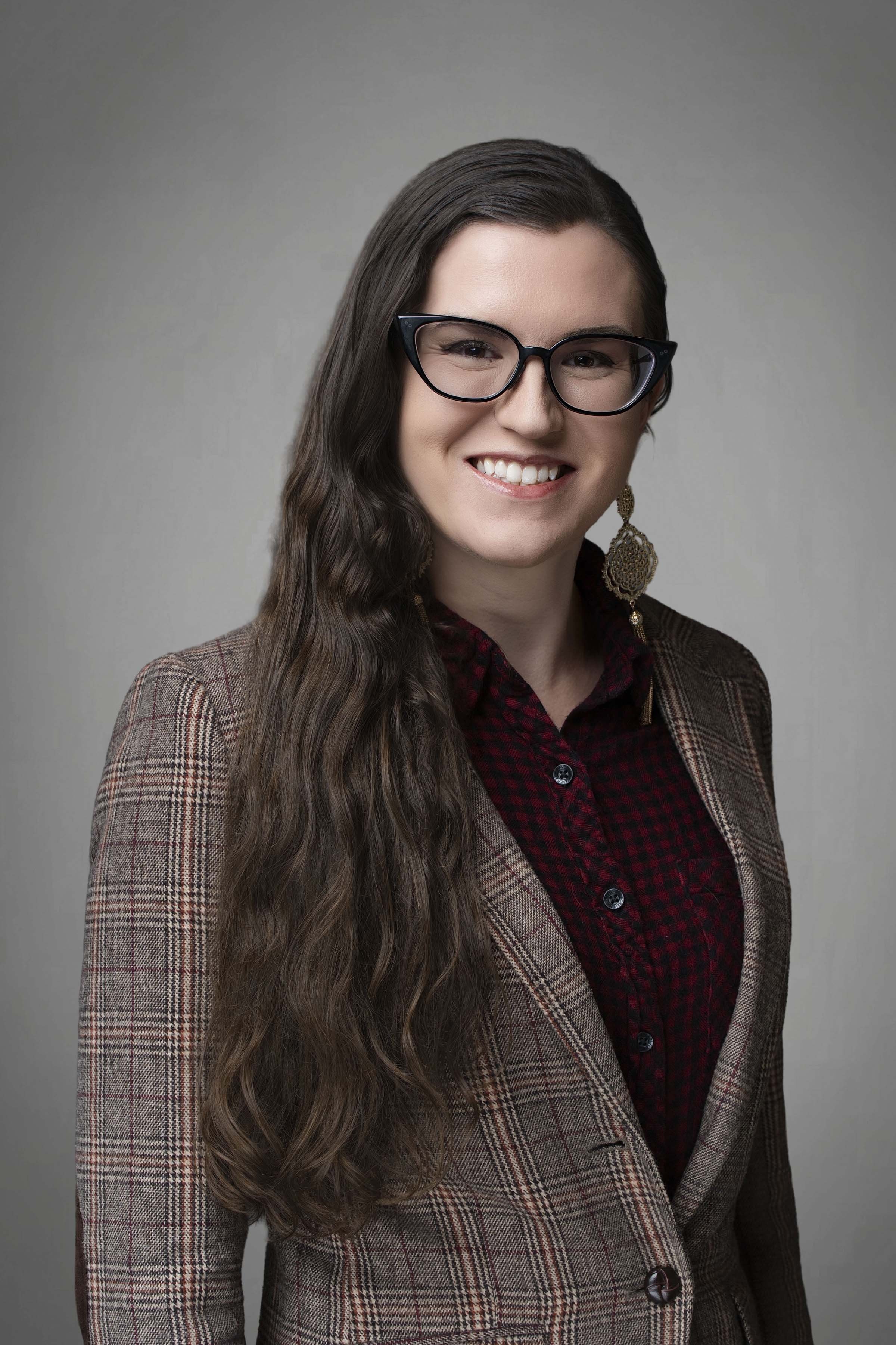 A woman with long wavy brown hair wearing glasses, a plaid blazer, and a red checkered shirt, smiling at the camera.