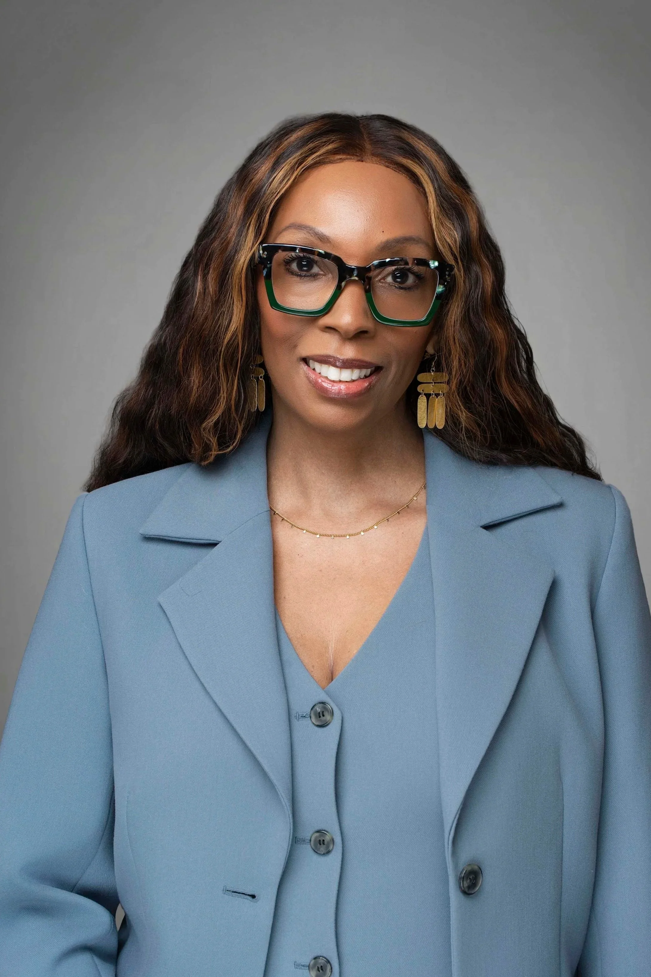 A woman with long wavy hair, wearing large black-rimmed glasses with green accents, a light blue blazer over a matching blouse, gold earrings, and a delicate gold necklace, smiling at the camera against a plain gray background.