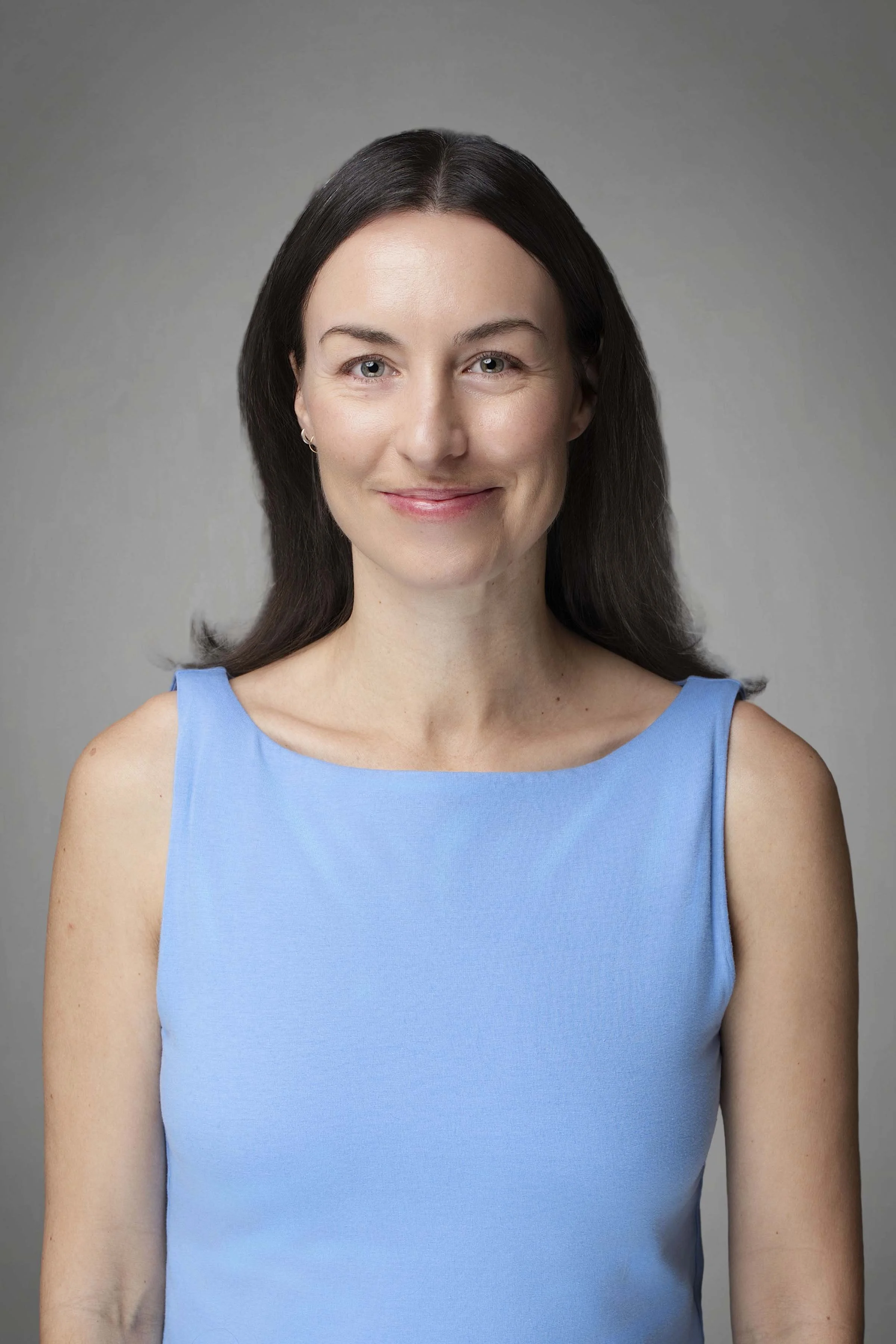 Headshot of a woman with fair skin, dark brown hair, and blue eyes, wearing a sleeveless light blue top and smiling gently against a neutral gray background.