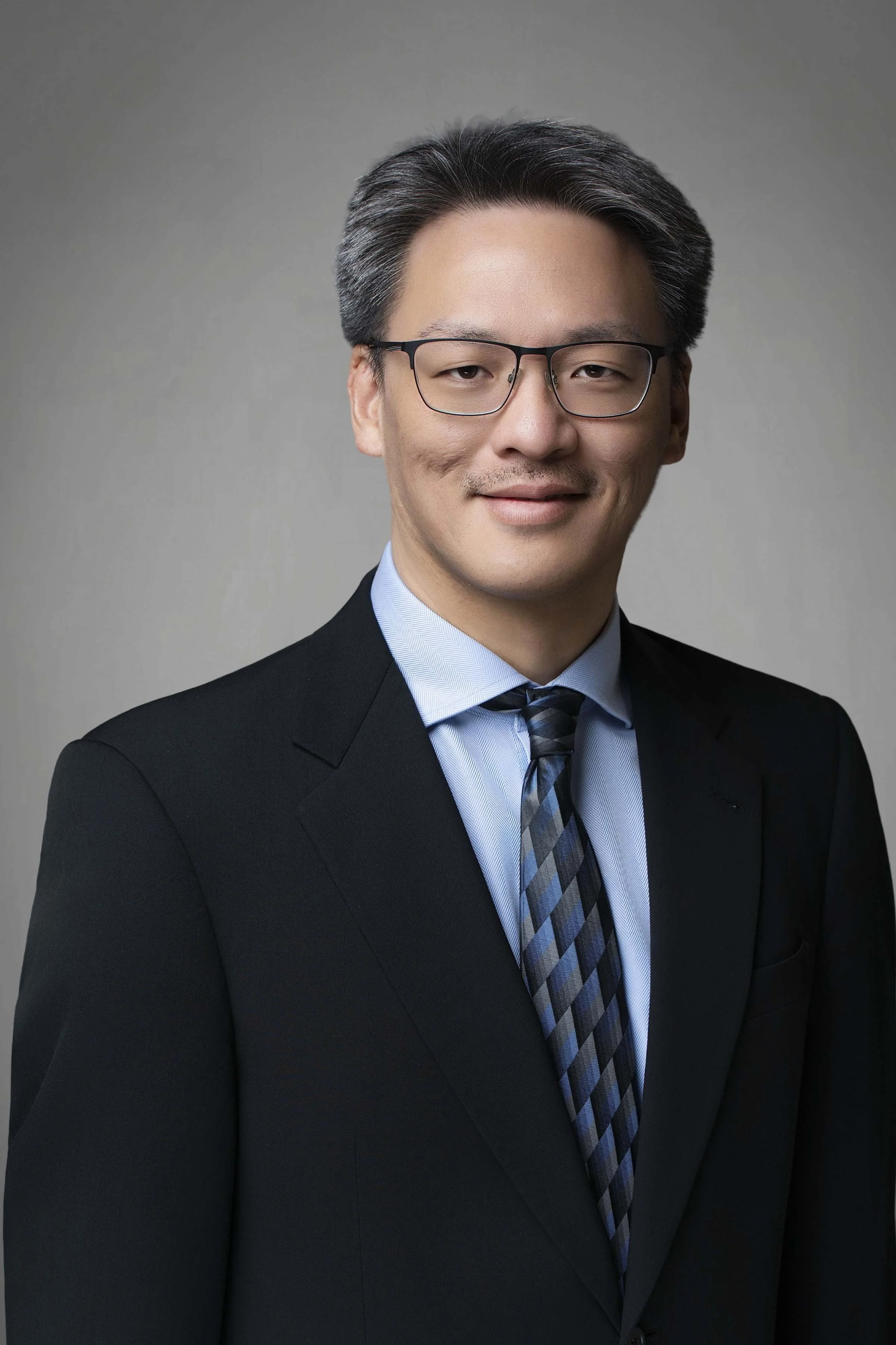 Professional portrait of an Asian man wearing glasses, a black suit, light blue shirt, and a blue plaid tie, smiling gently against a neutral background.