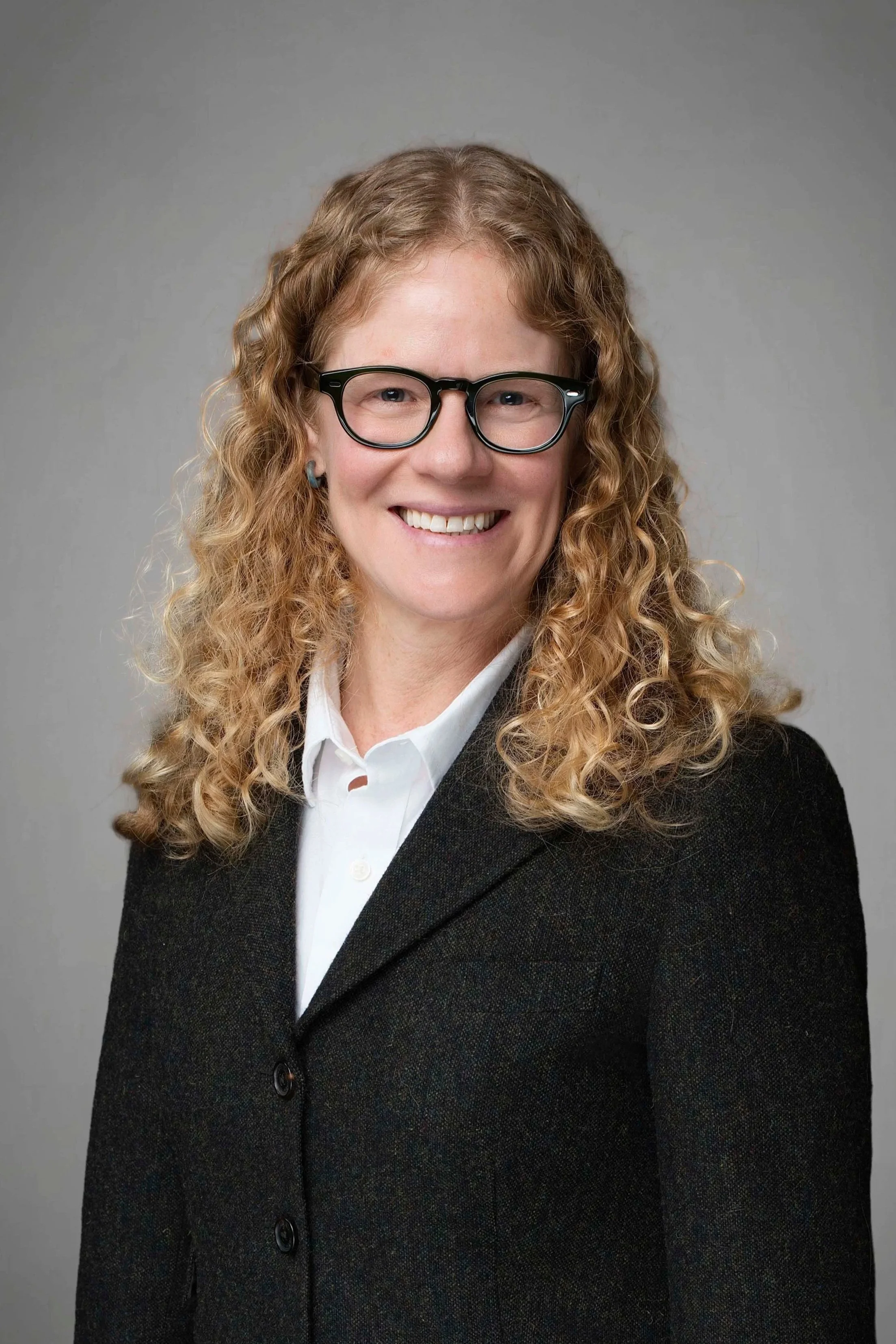 Professional woman with curly blonde hair, glasses, and a friendly smile, dressed in a black blazer and white shirt, against a gray background.