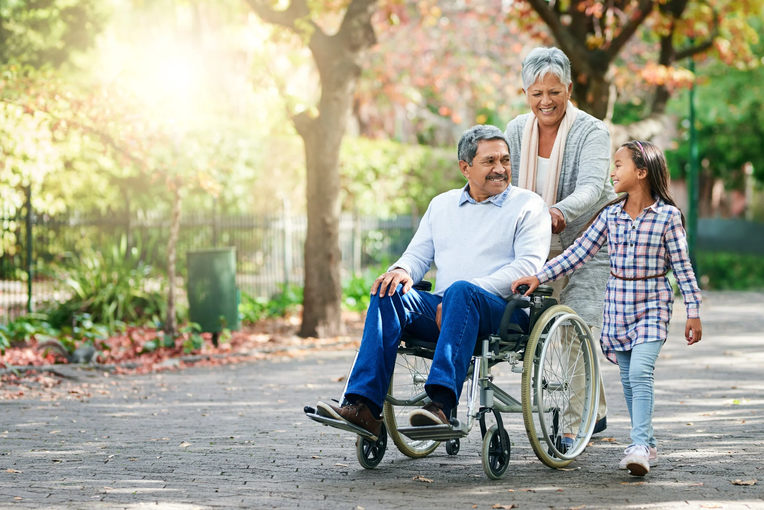 An elderly couple enjoys a walk in the park with their granddaughter, with the grandfather in a wheelchair.