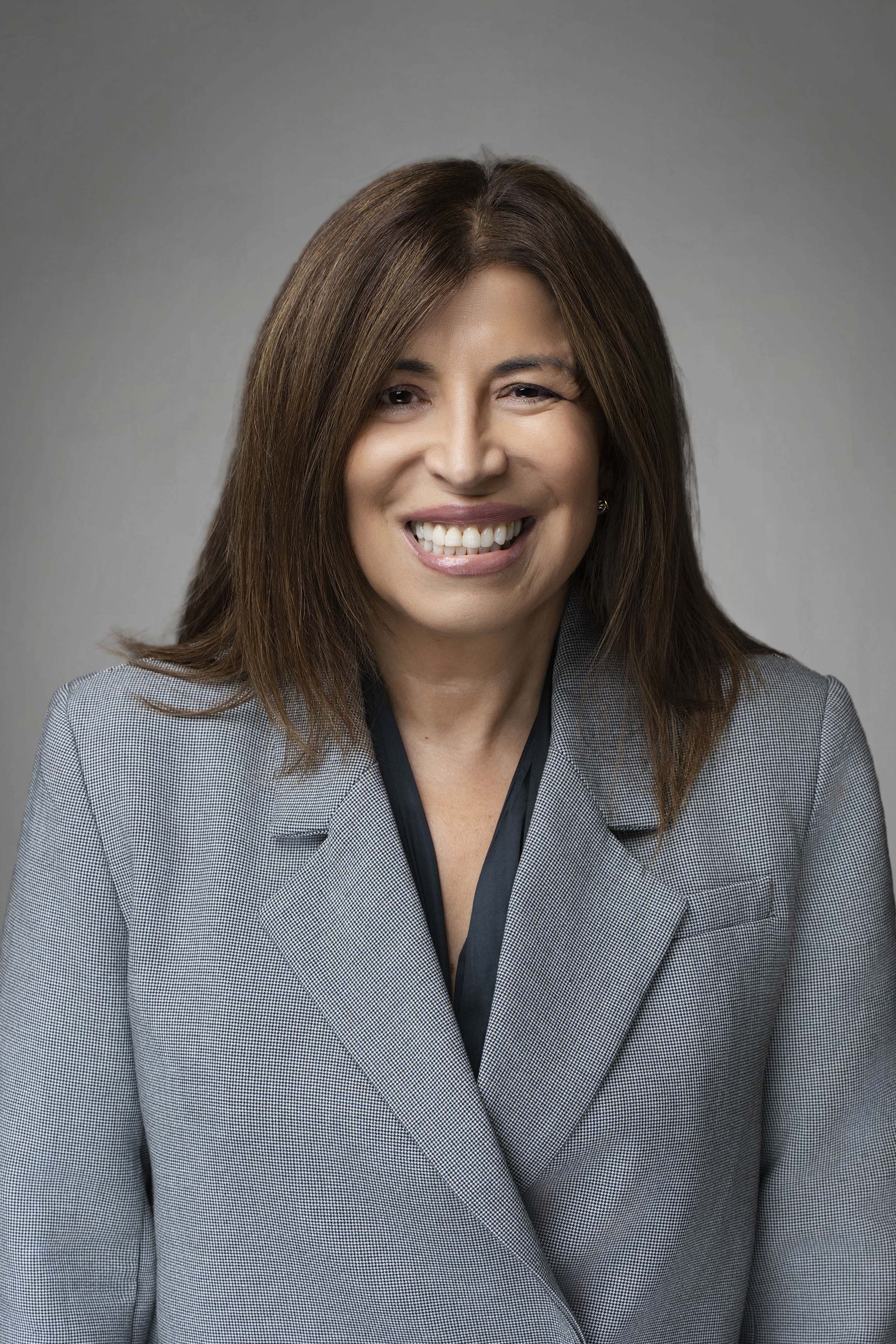 A professional woman with brown hair smiling, wearing a gray blazer and black top, against a gray background.