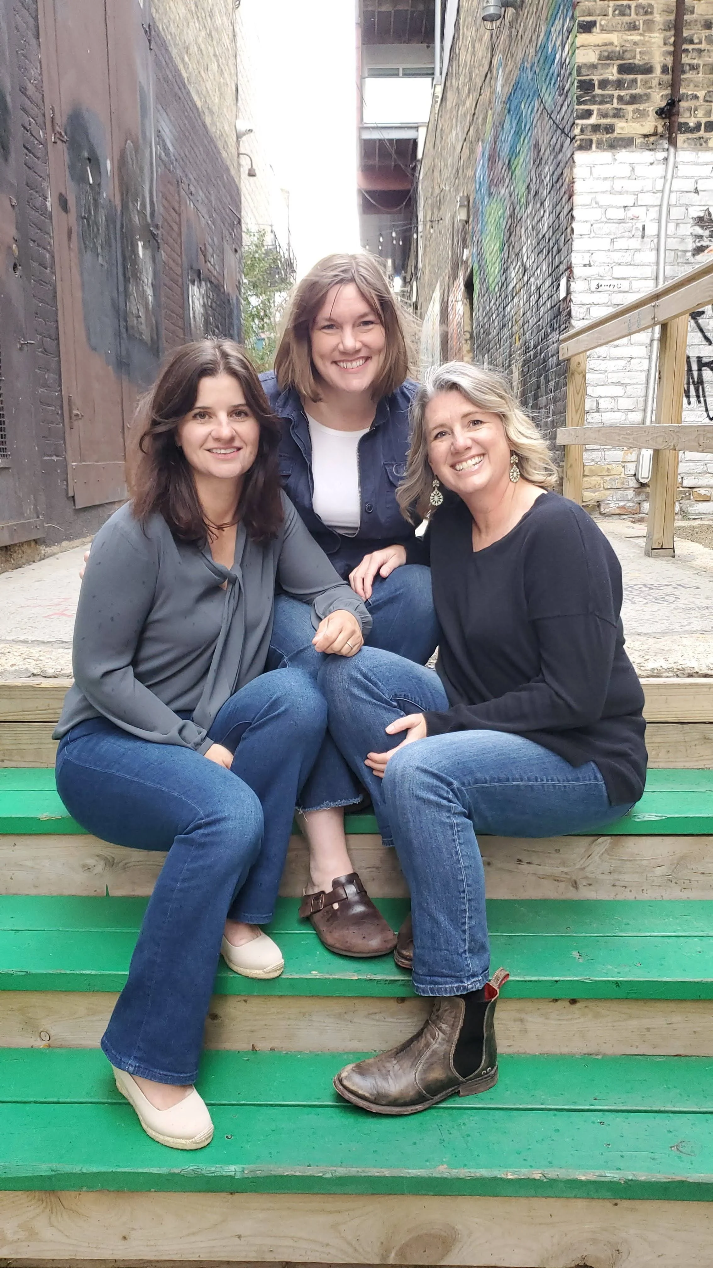 Three women sitting together outdoors on green wooden steps, smiling at the camera, in an alley with graffiti on brick walls.