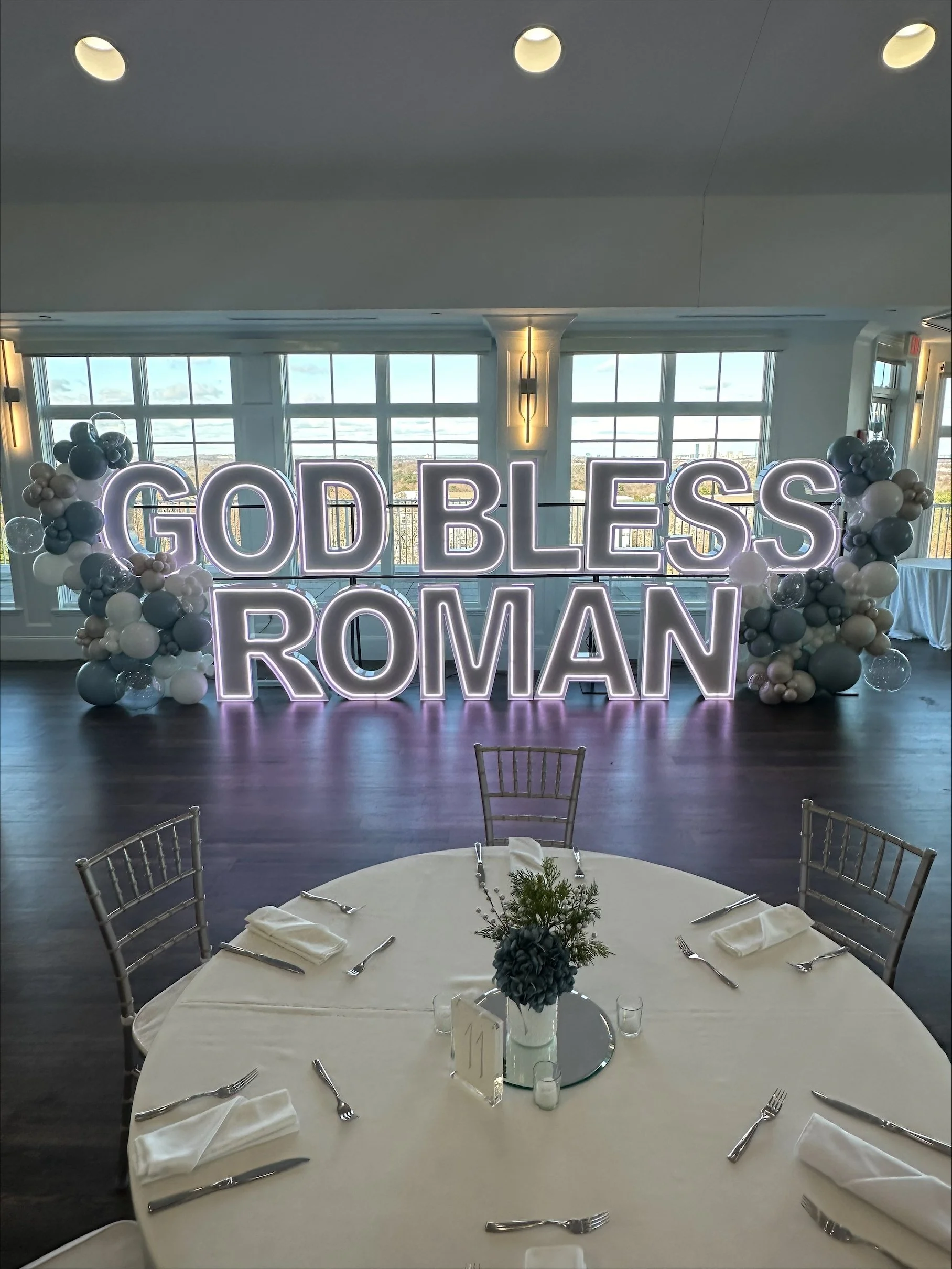 Decorative event with illuminated sign reading 'GOD BLESS ROMAN' behind a table set for a meal, with balloons surrounding the sign.