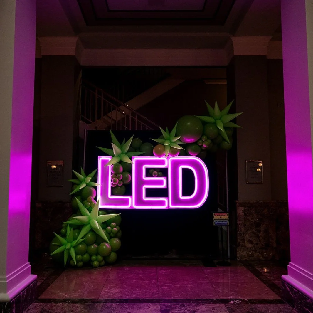 Pink neon sign spelling 'LED' surrounded by green balloon arrangements resembling succulents situated in an indoor setting.