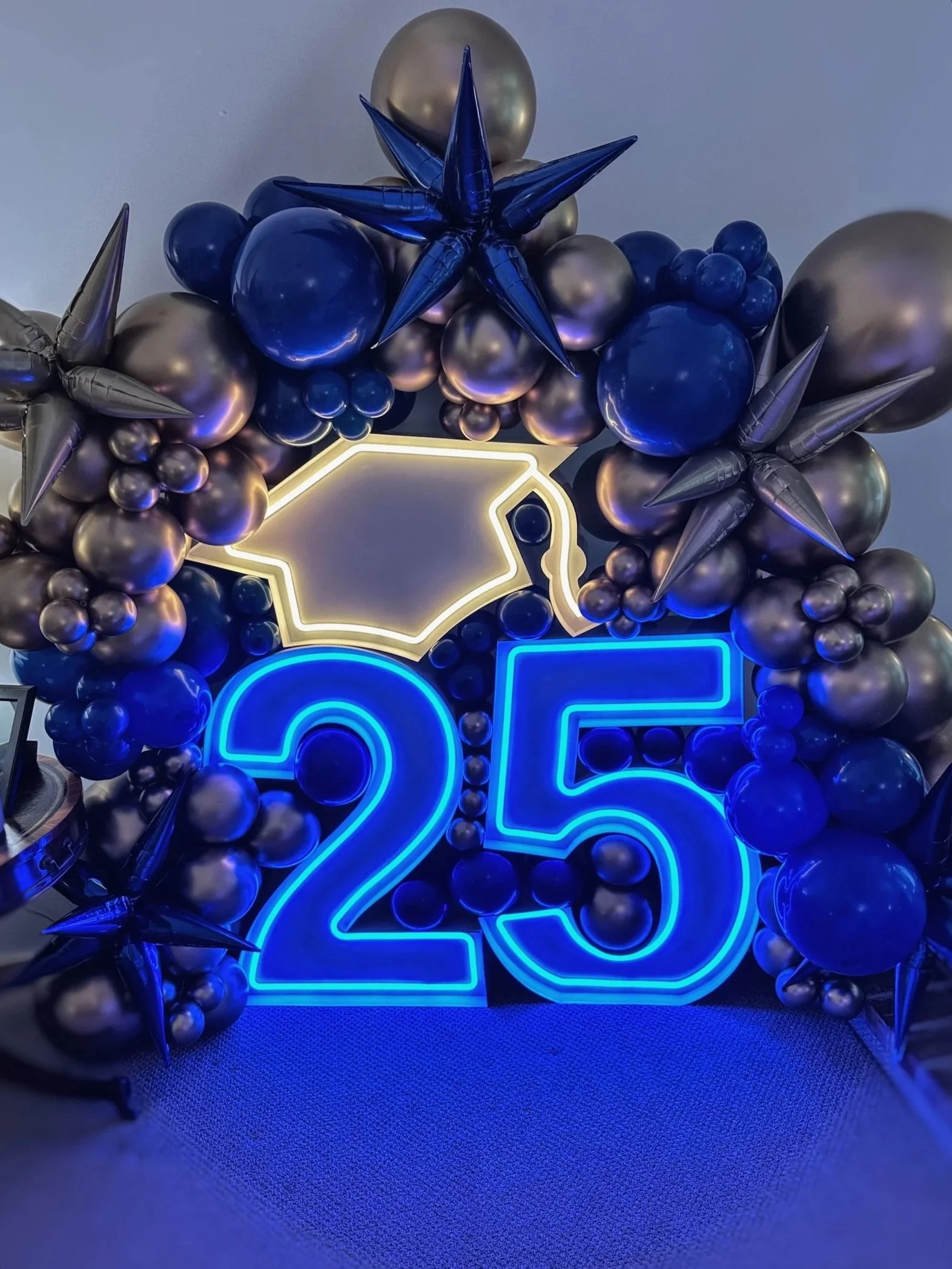 led marquees Neon sign reading '25' surrounded by metallic blue, purple, and silver balloons, with several star-shaped balloons managing some of the balloons, forming a colorful graduation decoration.
