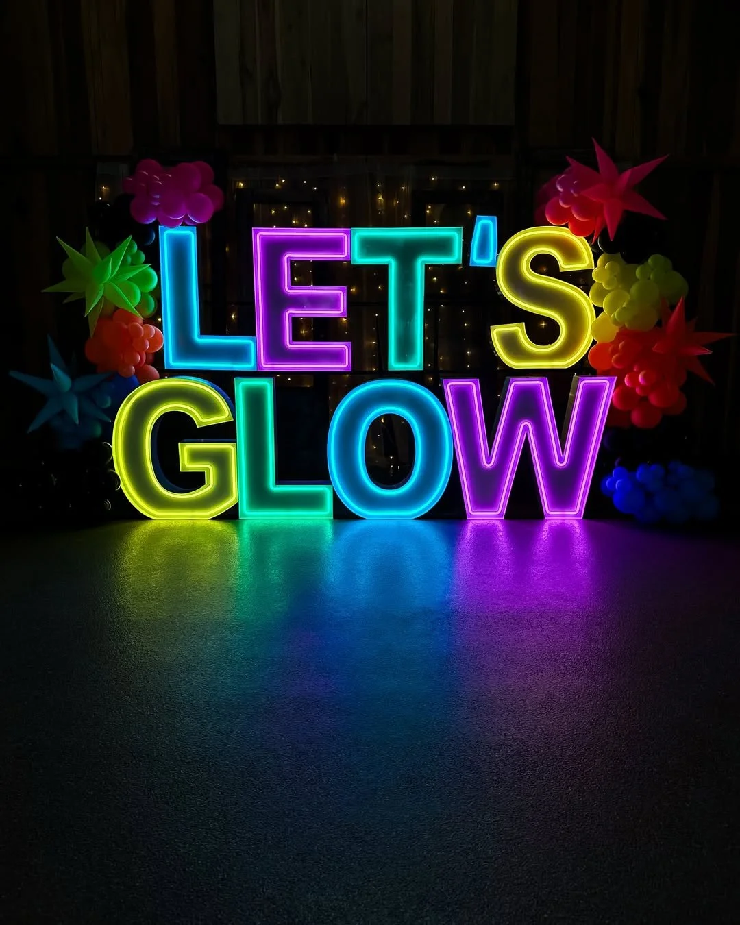 Neon sign reading 'LET'S GLOW' with colorful balloon and paper flower decorations in a dark setting.