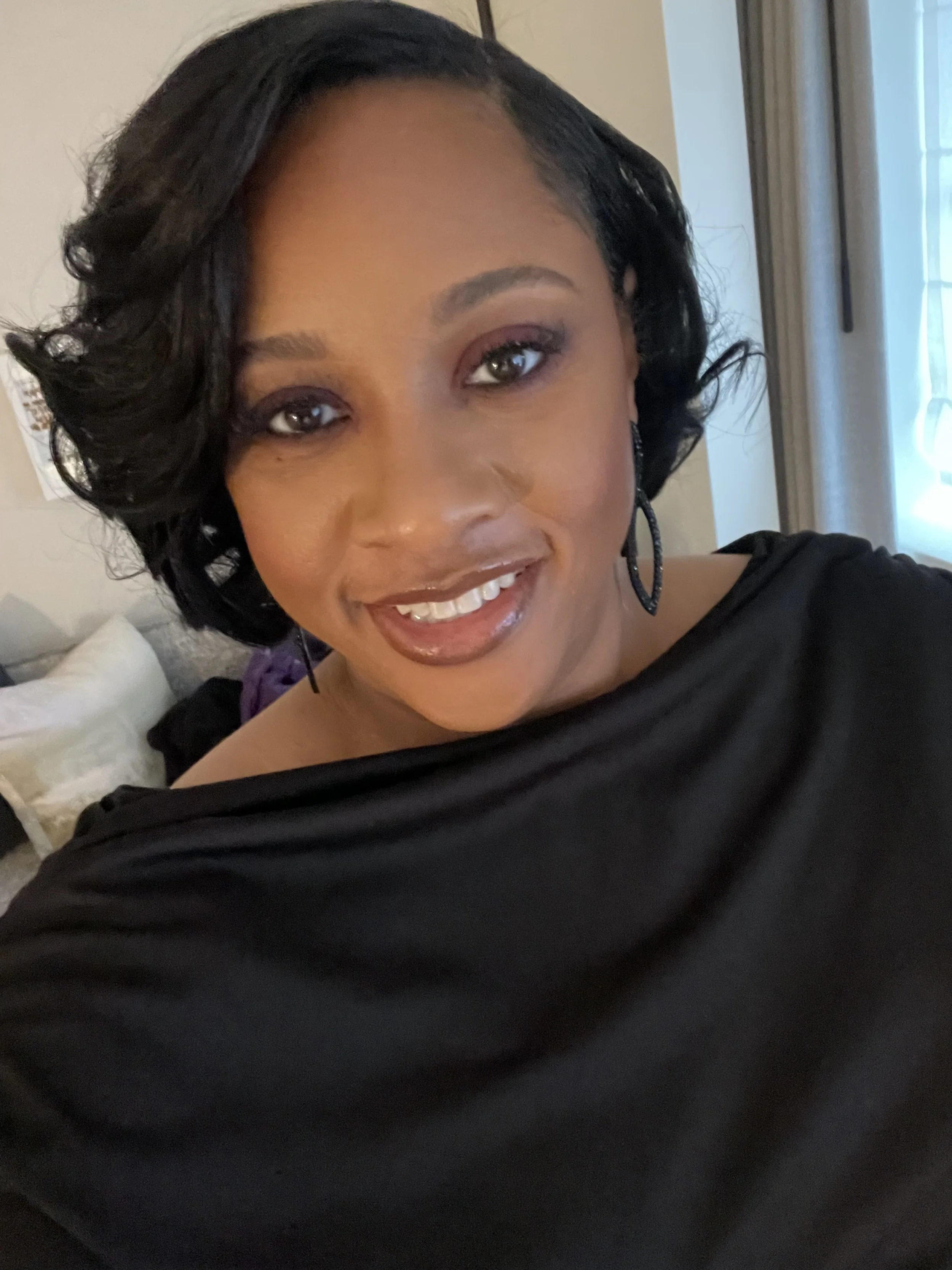 A woman with short, curly black hair, wearing earrings and a black top, smiling at the camera inside a room with beige walls and curtains.