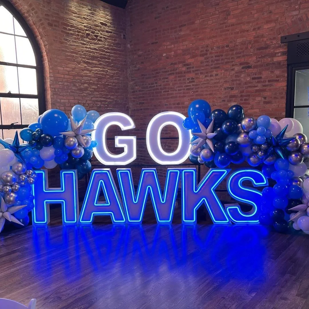 Neon sign that reads "GO HAWKS" with blue and white balloons and star-shaped decorations on each side, set against a brick wall, with wooden floor reflections.