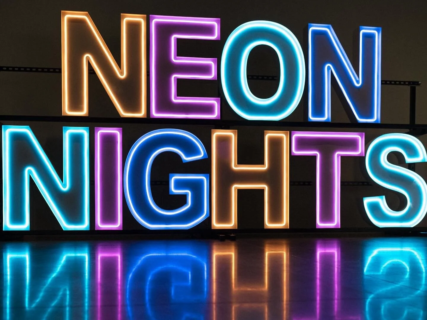 Neon sign spelling 'NEON NIGHTS' in colorful illuminated letters with reflections on the surface below.