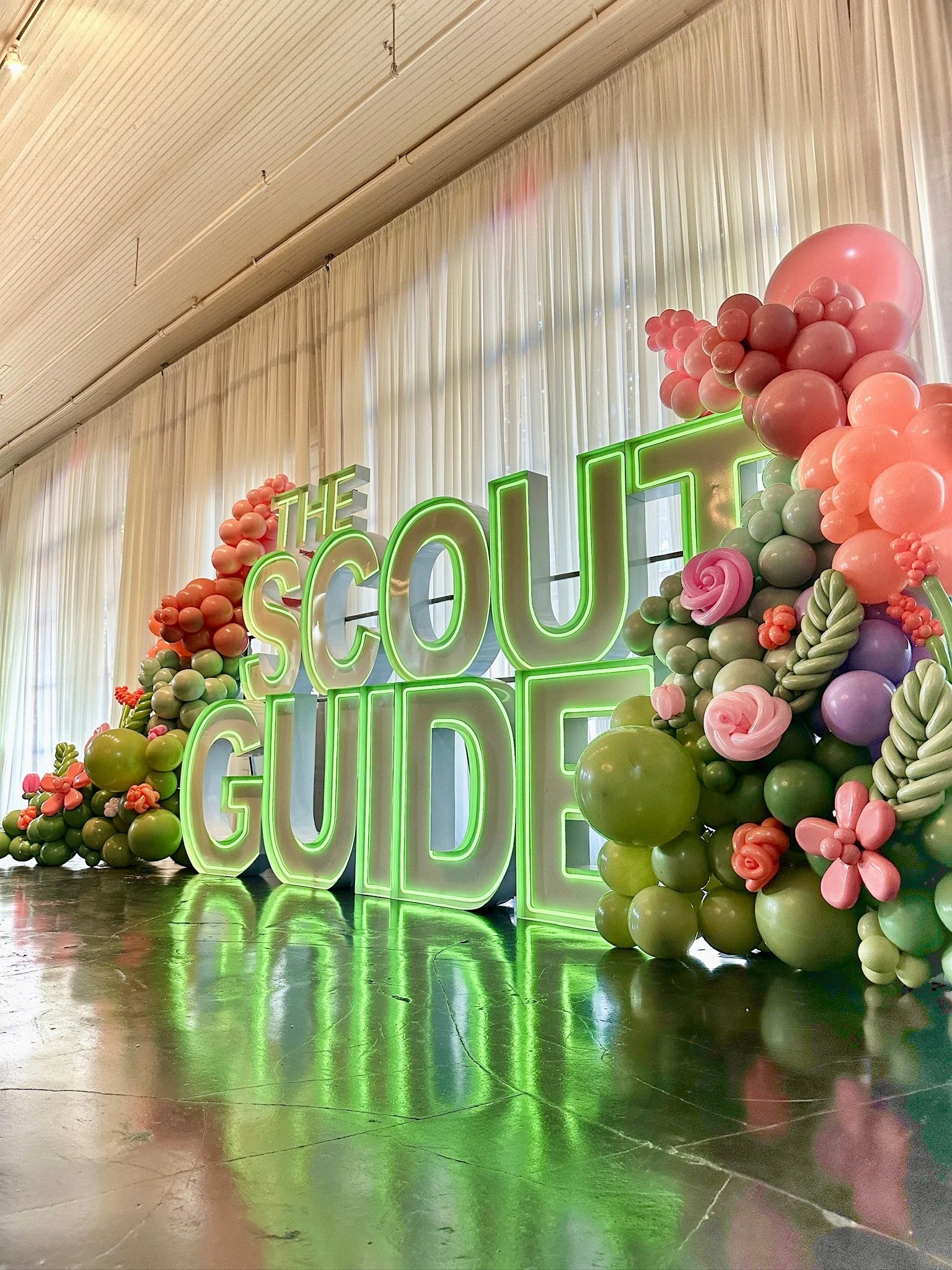 Neon sign reading 'The Scout Guide' with colorful balloon arrangements on either side, including pink, green, purple, and peach balloons, in an indoor setting with draped white curtains and a polished reflective floor.