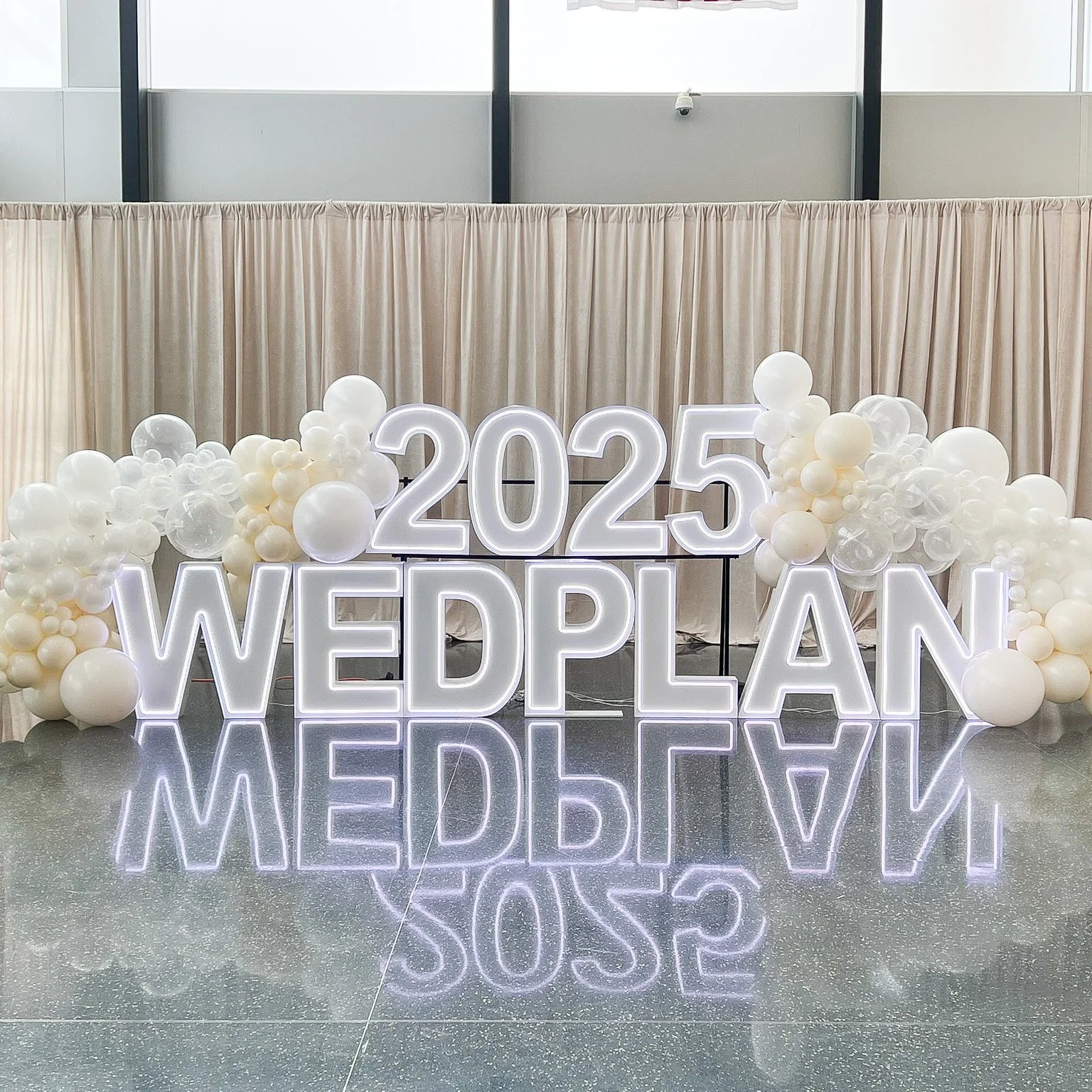 Lighting display with large white neon letters spelling '2025 WED PLAN' surrounded by white and transparent balloons, with a beige curtain backdrop and a mirror-like floor reflecting the display.