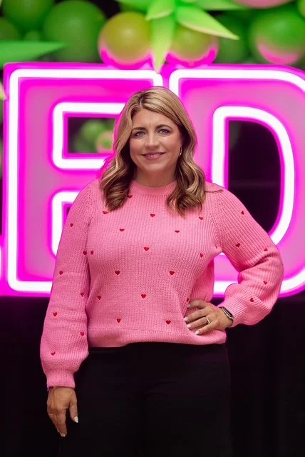A woman with blonde hair wearing a pink sweater with small red hearts standing in front of a neon pink and purple sign with green and purple balloons in the background.