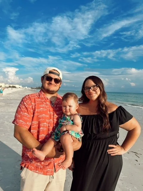 A family of three at the beach with the ocean and blue sky in the background. The man is wearing sunglasses and a red patterned shirt, holding a young girl in a colorful dress, while the woman in a black dress and glasses stands beside them.