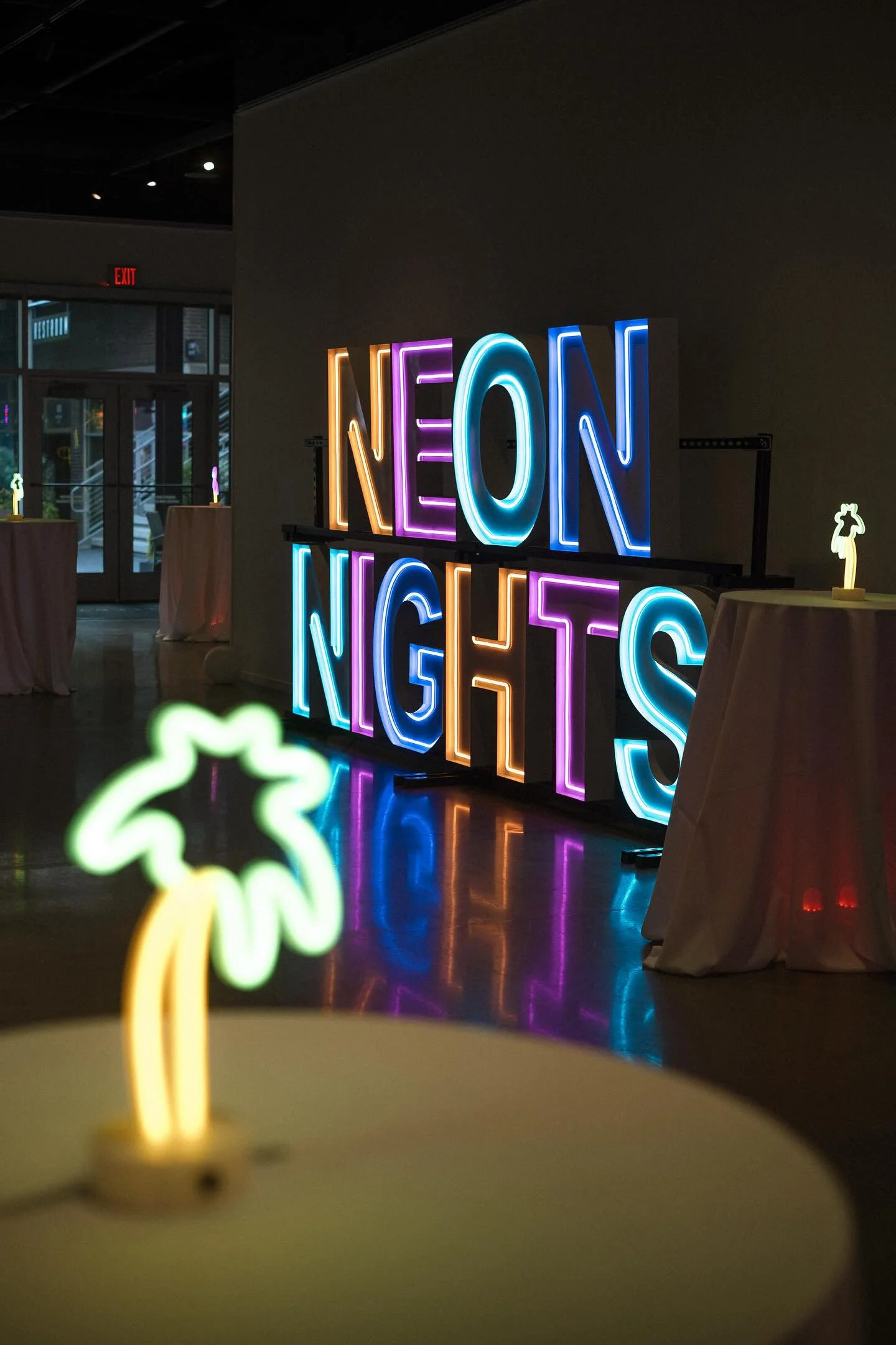 Colorful neon sign reading 'NEON NIGHTS' in a dark indoor space with small decorative palm trees on tables.
