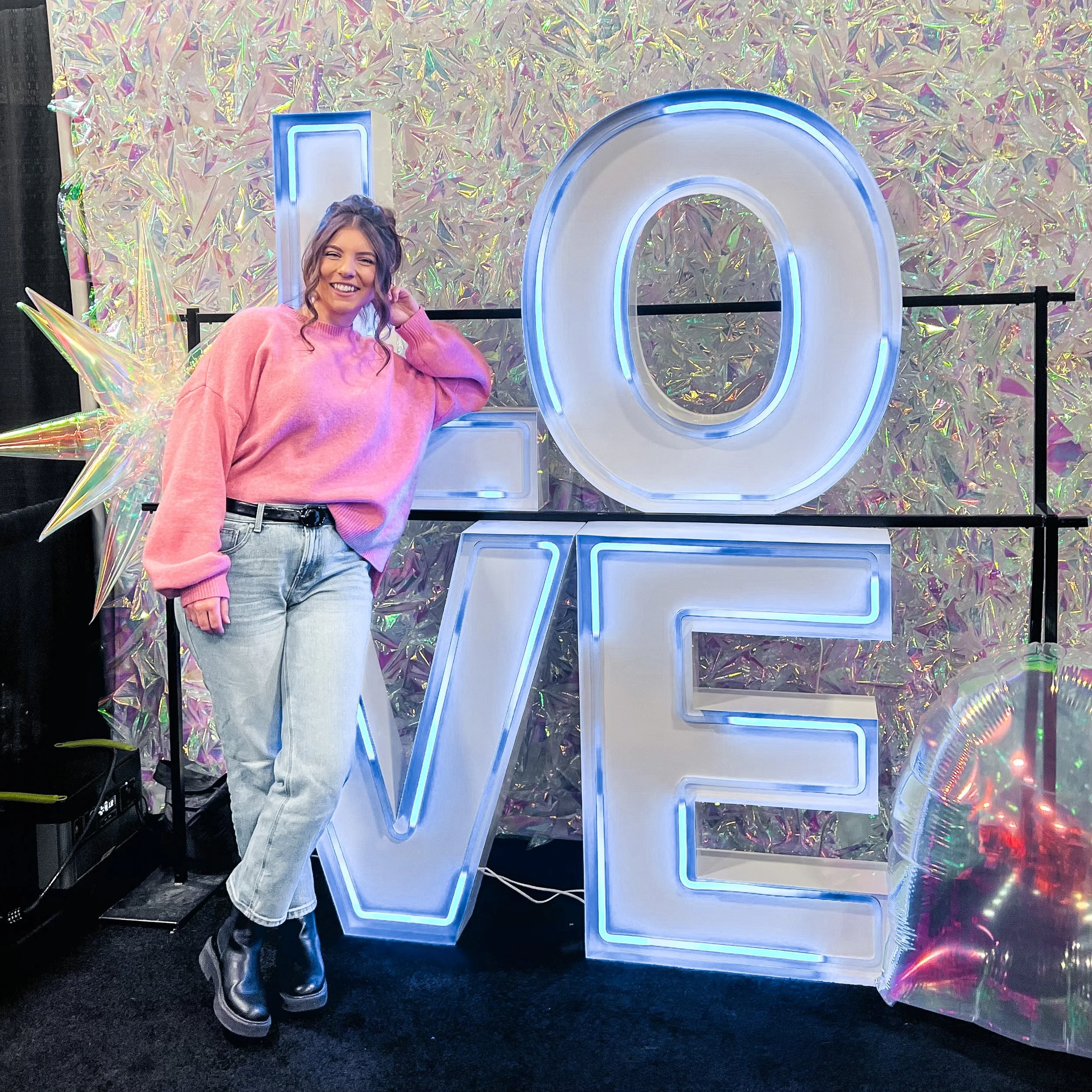 A woman dressed in a pink oversized sweater, light jeans, and black boots poses next to a neon sign that says 'LOVE' with large illuminated letters, set against a holographic background.