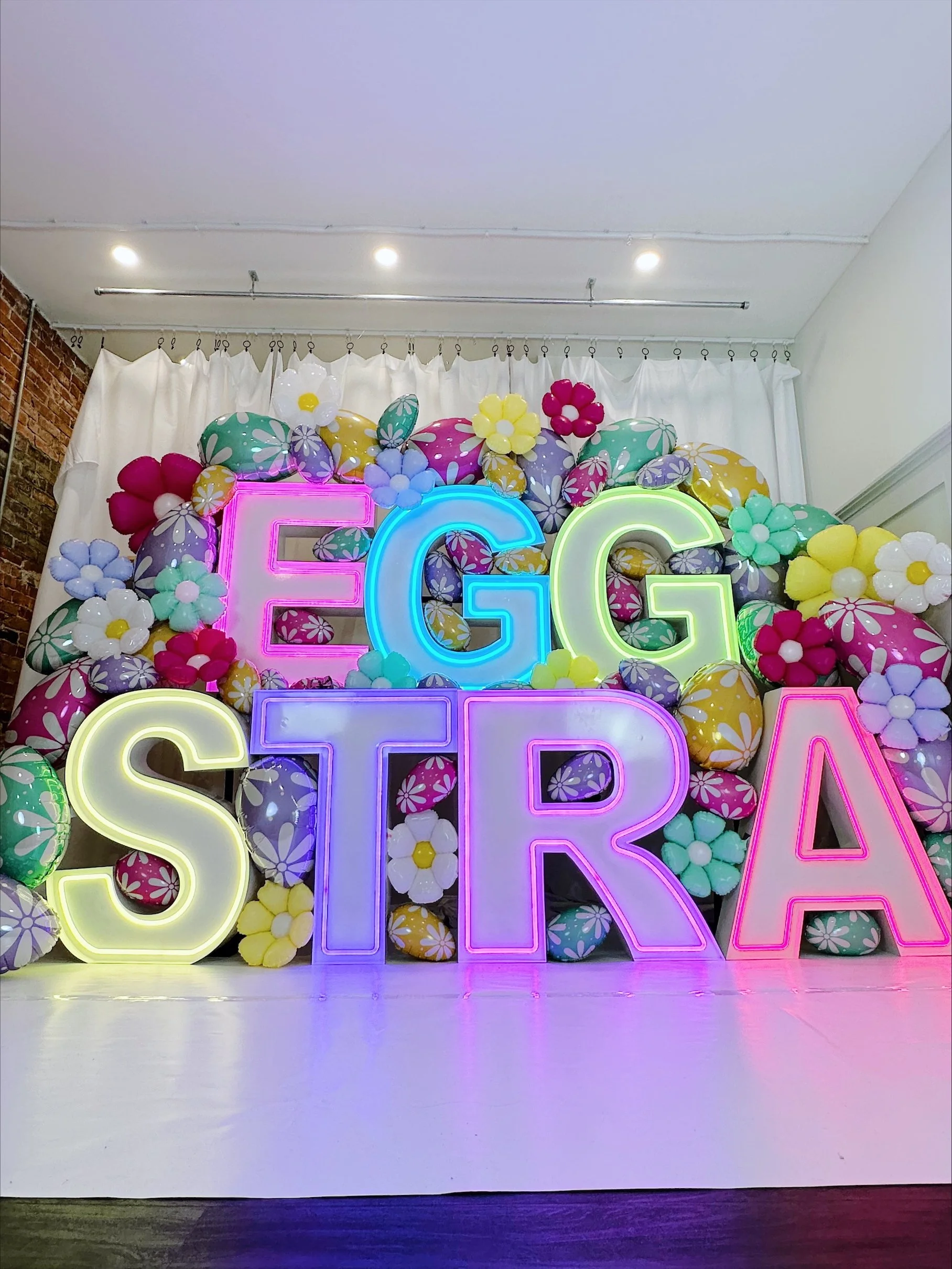 Colorful Easter neon led letters spelling 'EGGSTRA' surrounded by balloons shaped like Easter eggs and flowers, set against a white curtain backdrop.