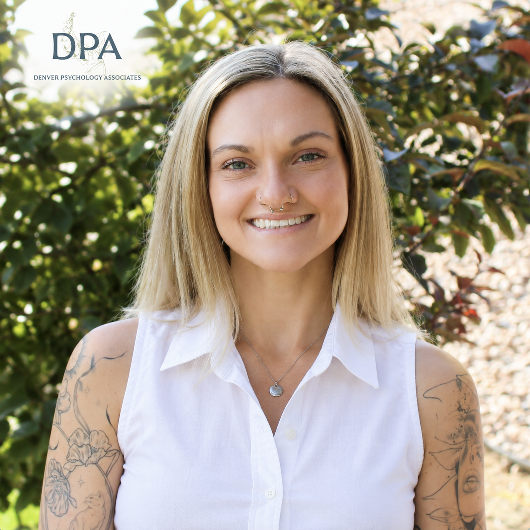 A young woman with blonde hair, tattoos, and a nose ring smiling outdoors with green foliage in the background.