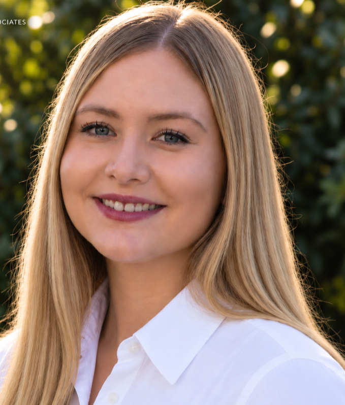 Close-up of a young woman with long blonde hair, smiling, wearing a white shirt, outdoors with greenery in the background.