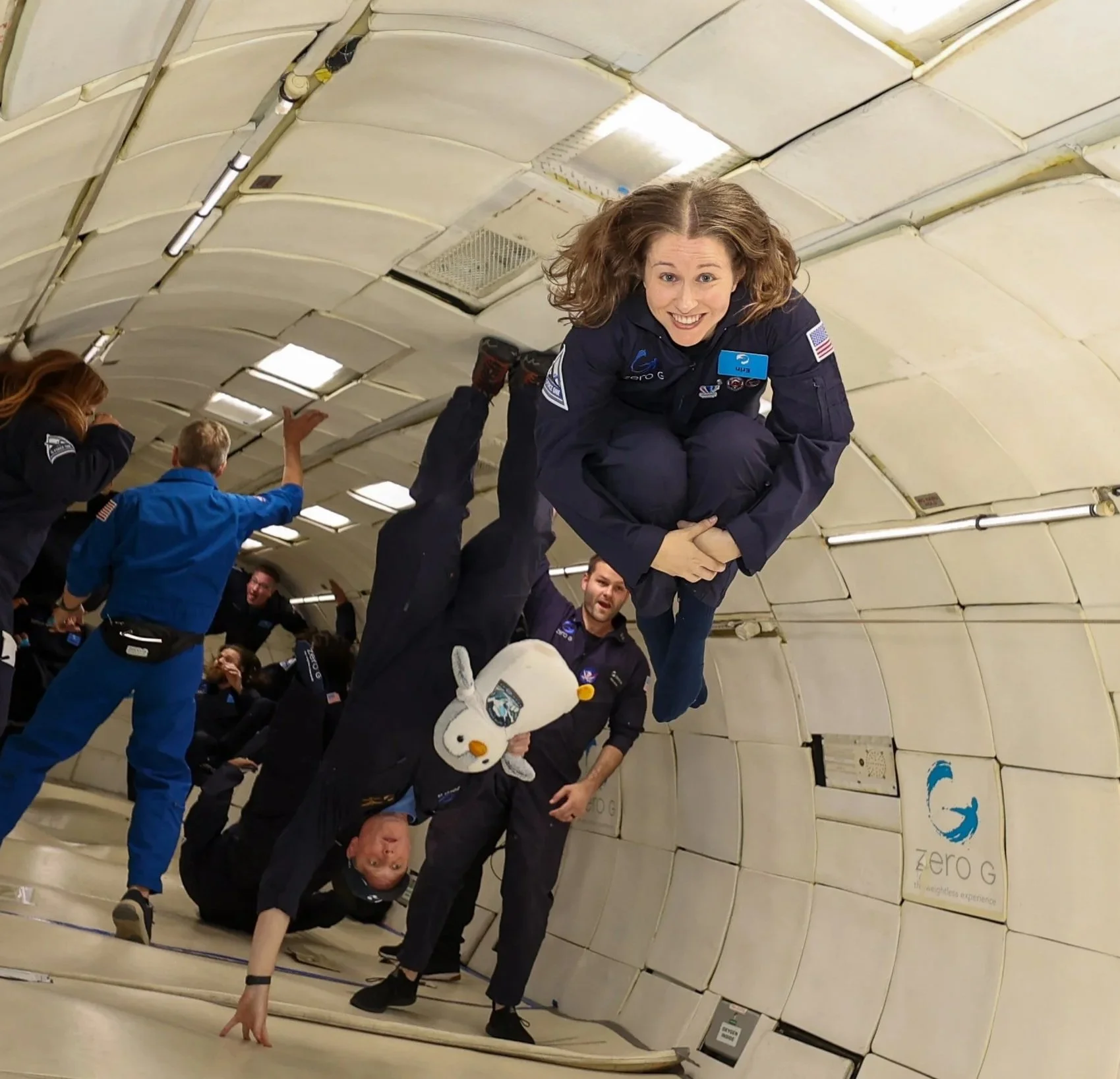 Astronauts inside a spacecraft, some posing and one hanging upside down, with the zero gravity sign visible in the background.