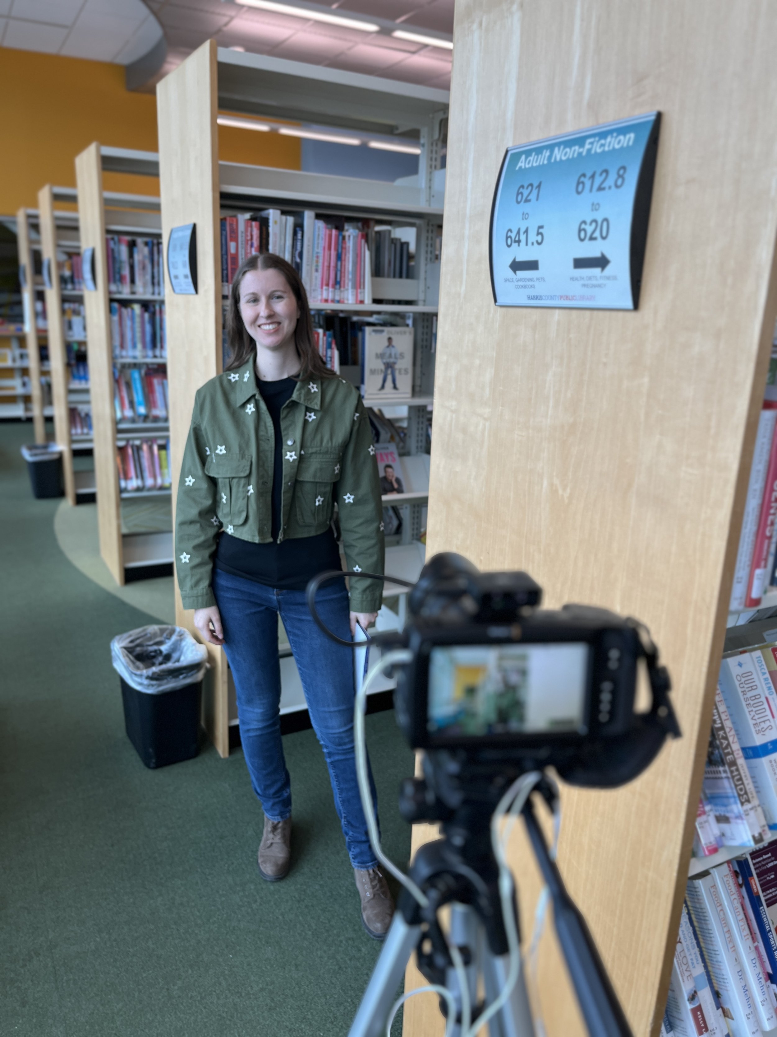 A woman smiling and standing in a library, facing a camera on a tripod, with bookshelves in the background.