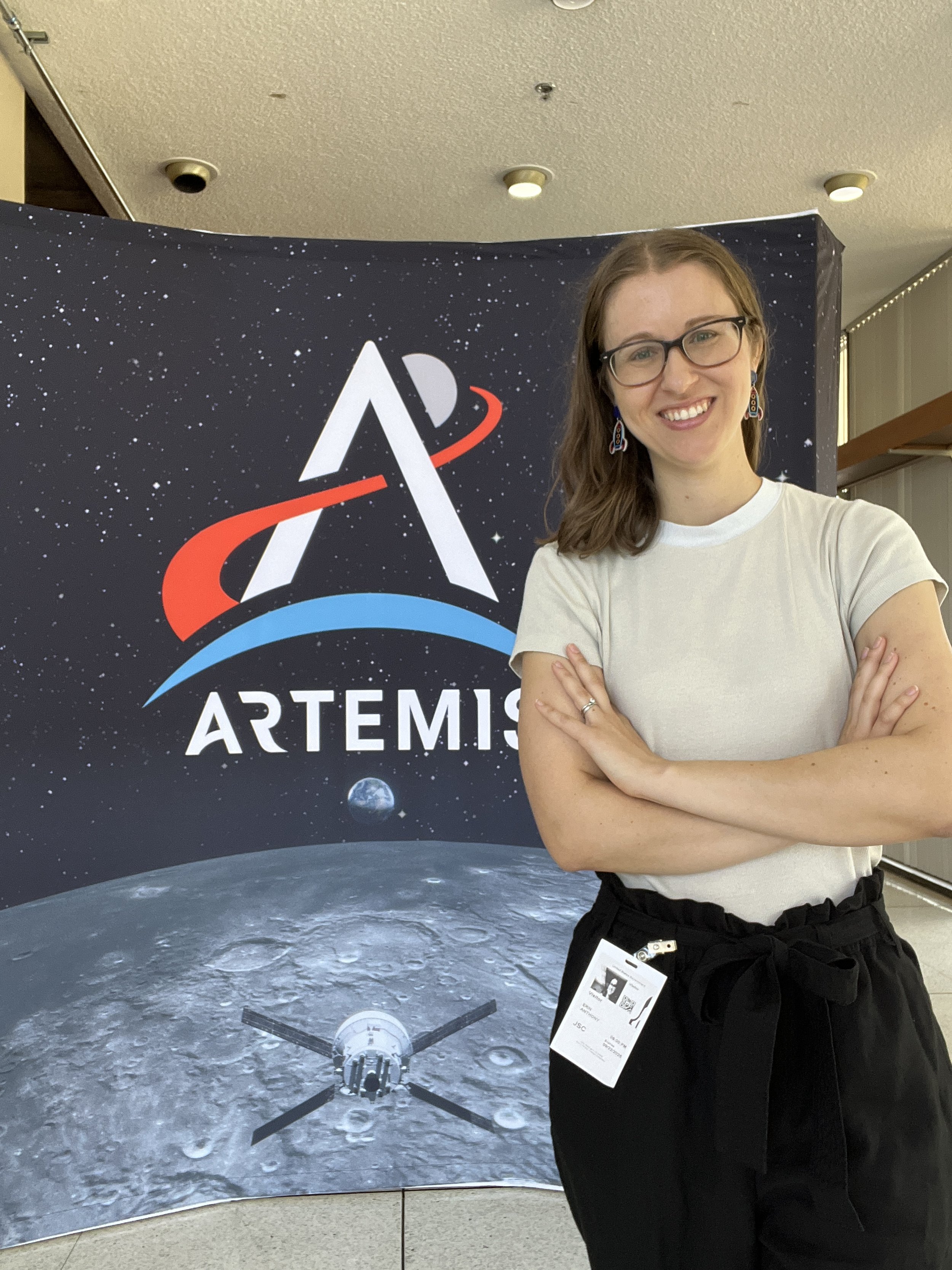 A smiling woman with glasses and earrings stands with crossed arms in front of a space-themed backdrop featuring the Artemis logo, the moon, and a spacecraft.