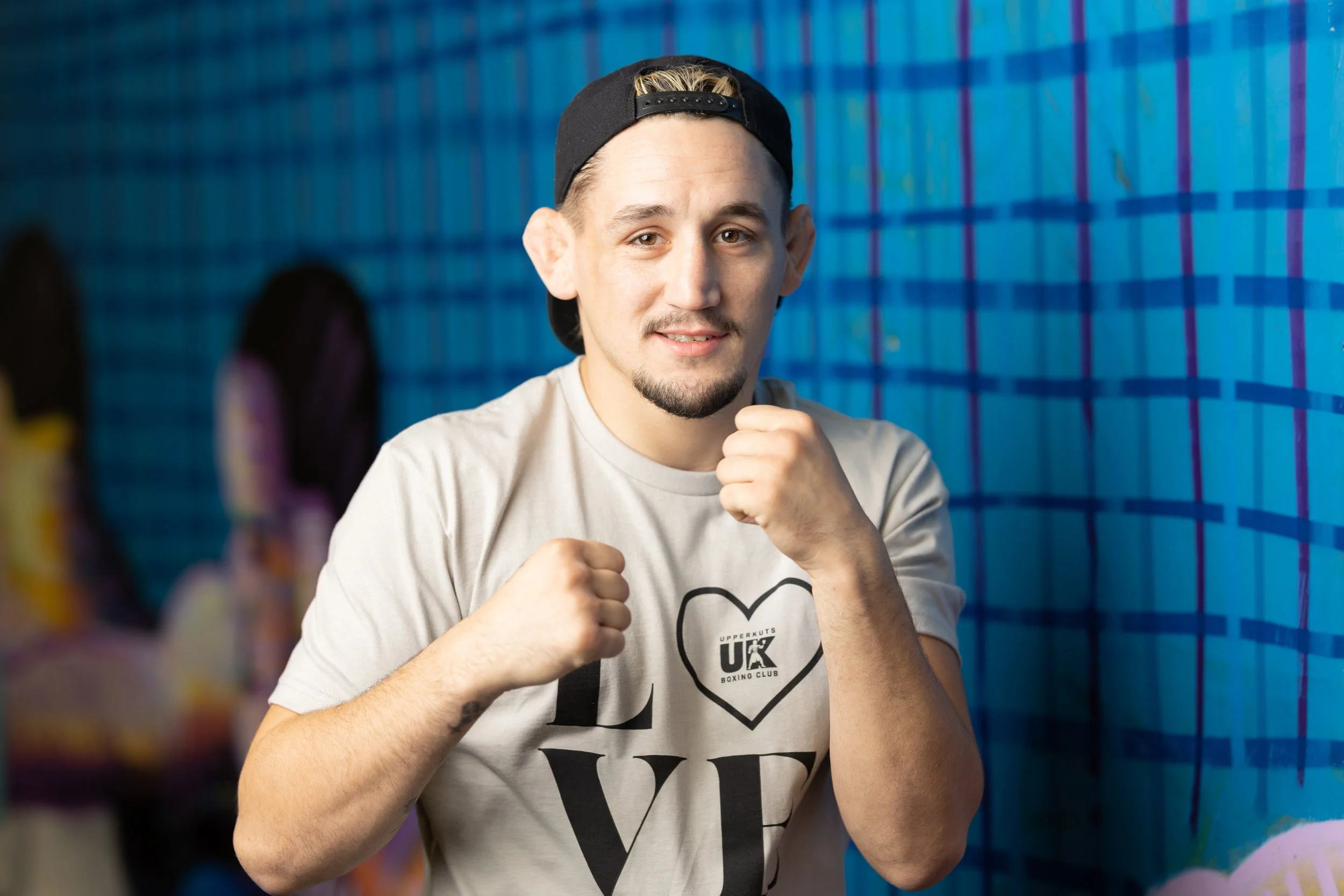 A young man wearing a white T-shirt with the word "LOVE" and a heart shape on it, posing with fists raised in a boxing stance, in front of a blue graffiti wall.