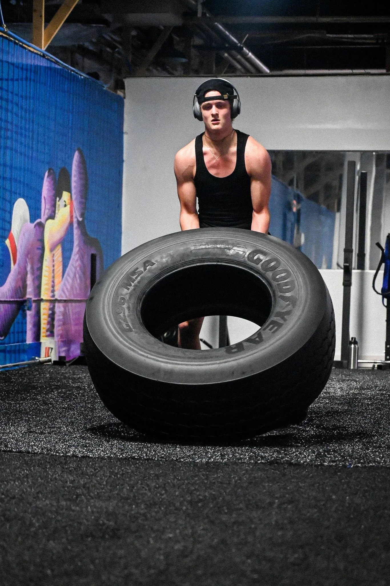 A young man lifting a large Goodyear tire in a gym with graffiti artwork on the wall.