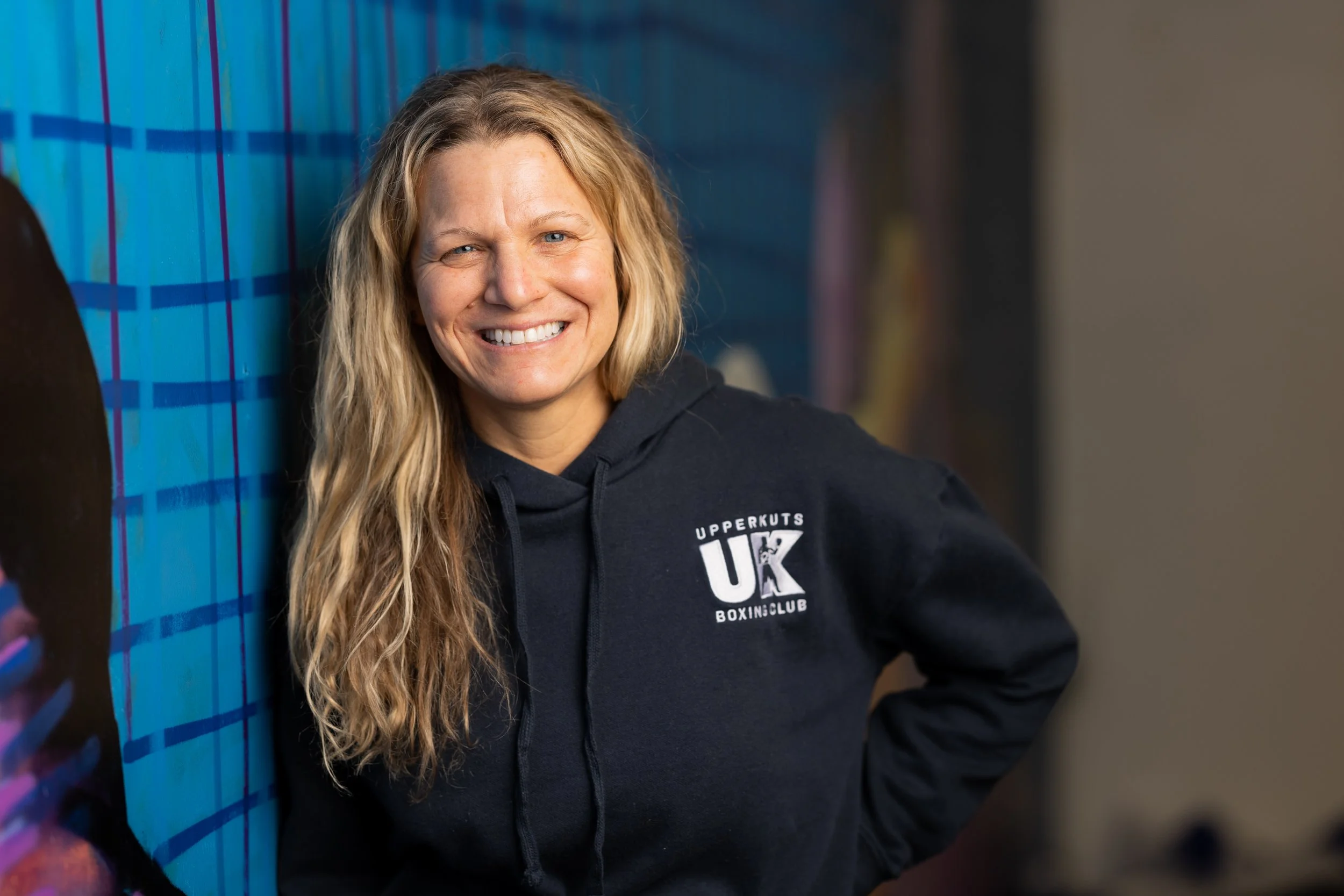 A smiling woman with long blonde hair, wearing a black hoodie with the logo for 'Uppercut's UK Boxing Club,' standing indoors against a blue wall.