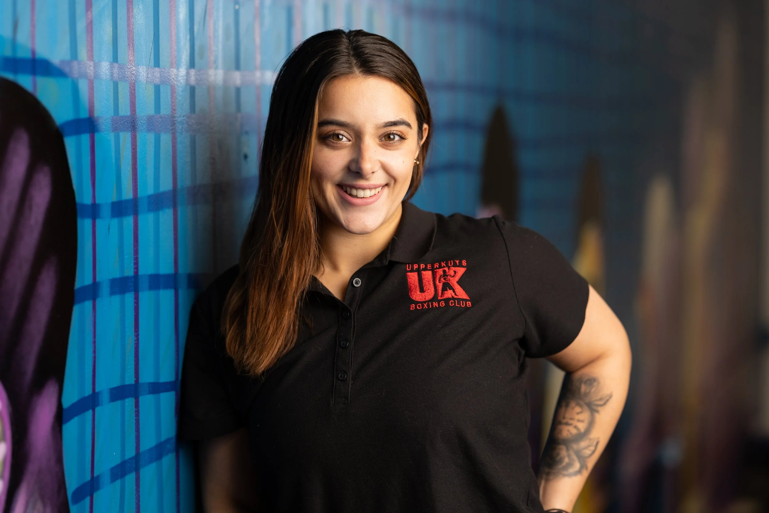 A young woman with long brown hair smiling and standing against a graffiti-covered wall wearing a black polo shirt with red text and logo for Uppercuts UK Boxing Club.