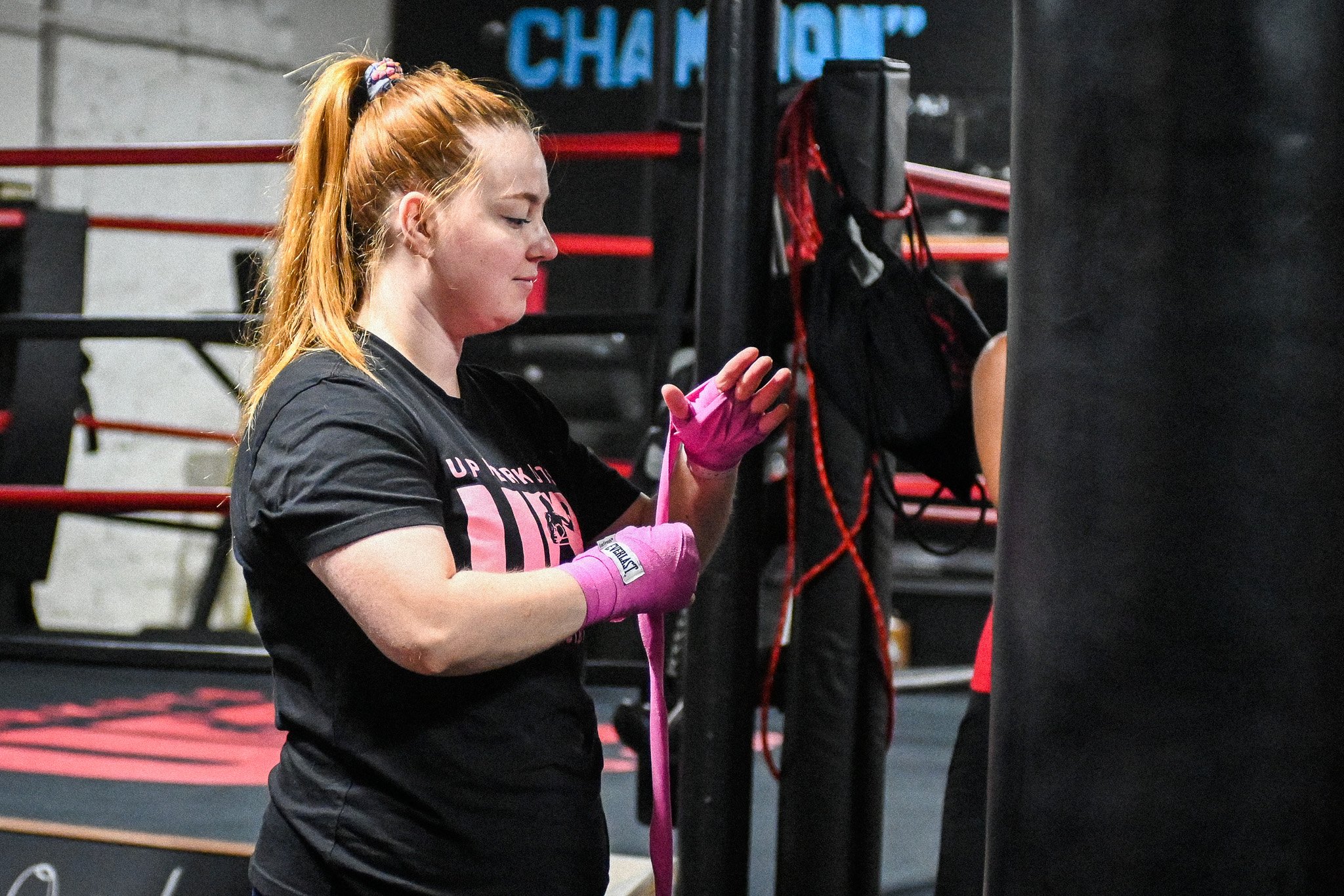 Red-haired woman in a black T-shirt and pink hand wraps, tying her boxing gloves in a gym.