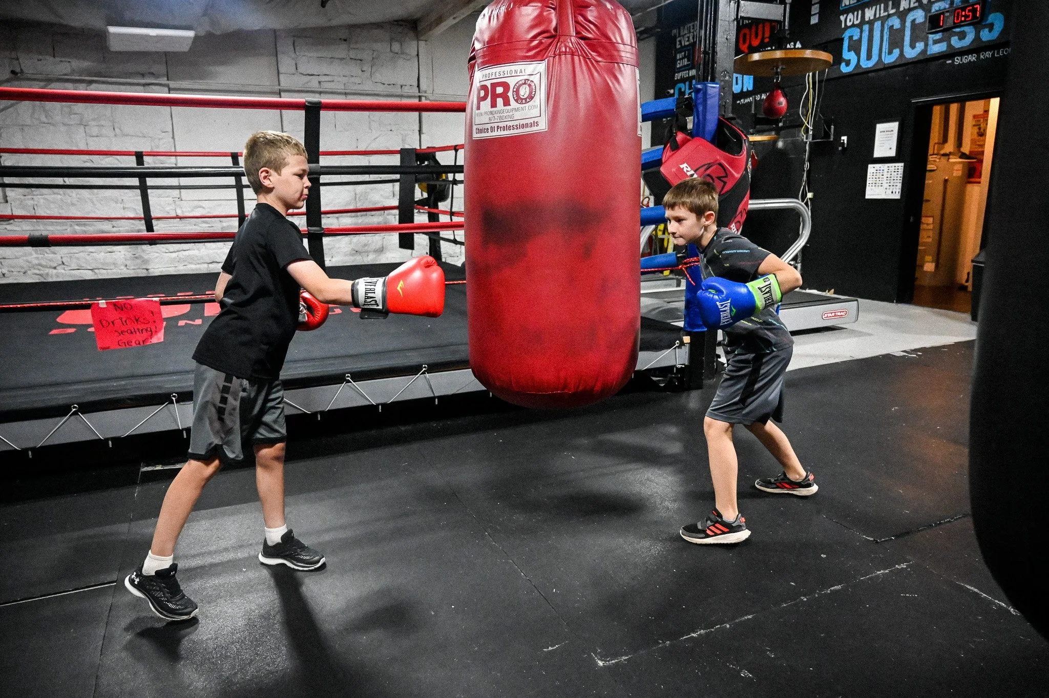 Two boys practicing boxing in a gym, facing each other with boxing gloves, near a red punching bag.