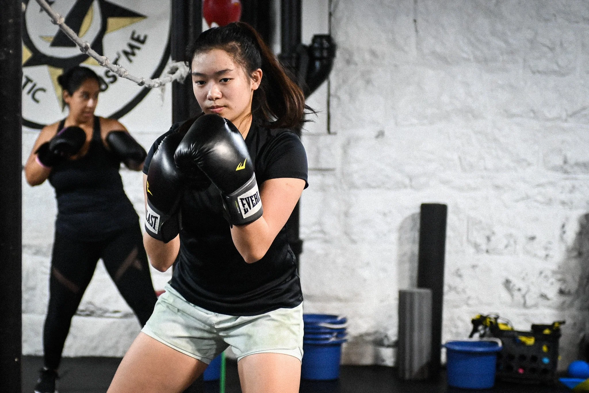 Two women practicing boxing in a gym. The woman in the foreground is wearing black boxing gloves and focused, with her hair tied back. The woman in the background is also in black, with her hands up in a boxing stance, and a wall with various equipment is visible behind them.