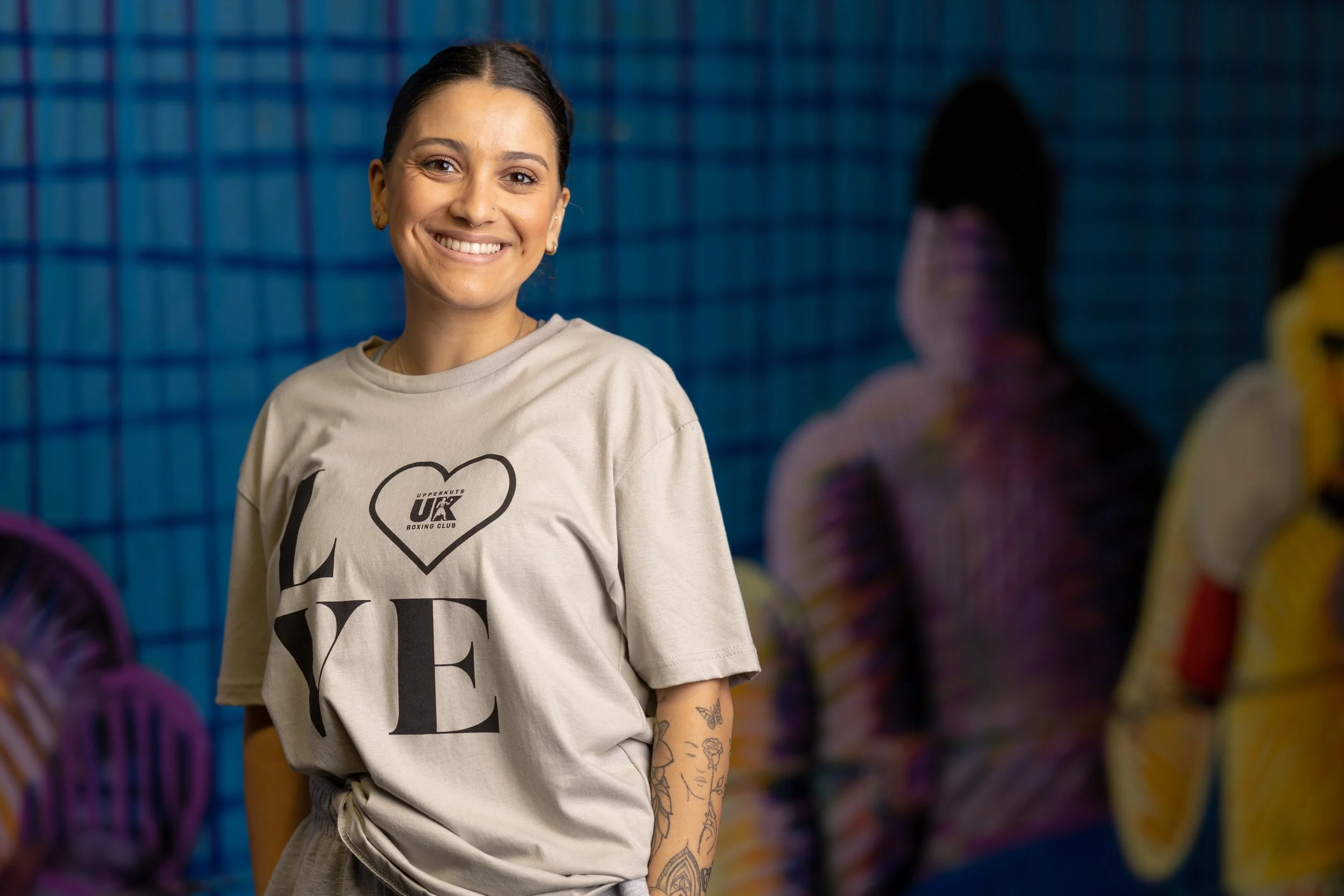 Smiling woman with dark hair and tattoos on her arm, wearing a beige T-shirt with the word 'LOVE' and a heart with 'UK' inside, standing indoors with a blurred background of people and colorful objects.