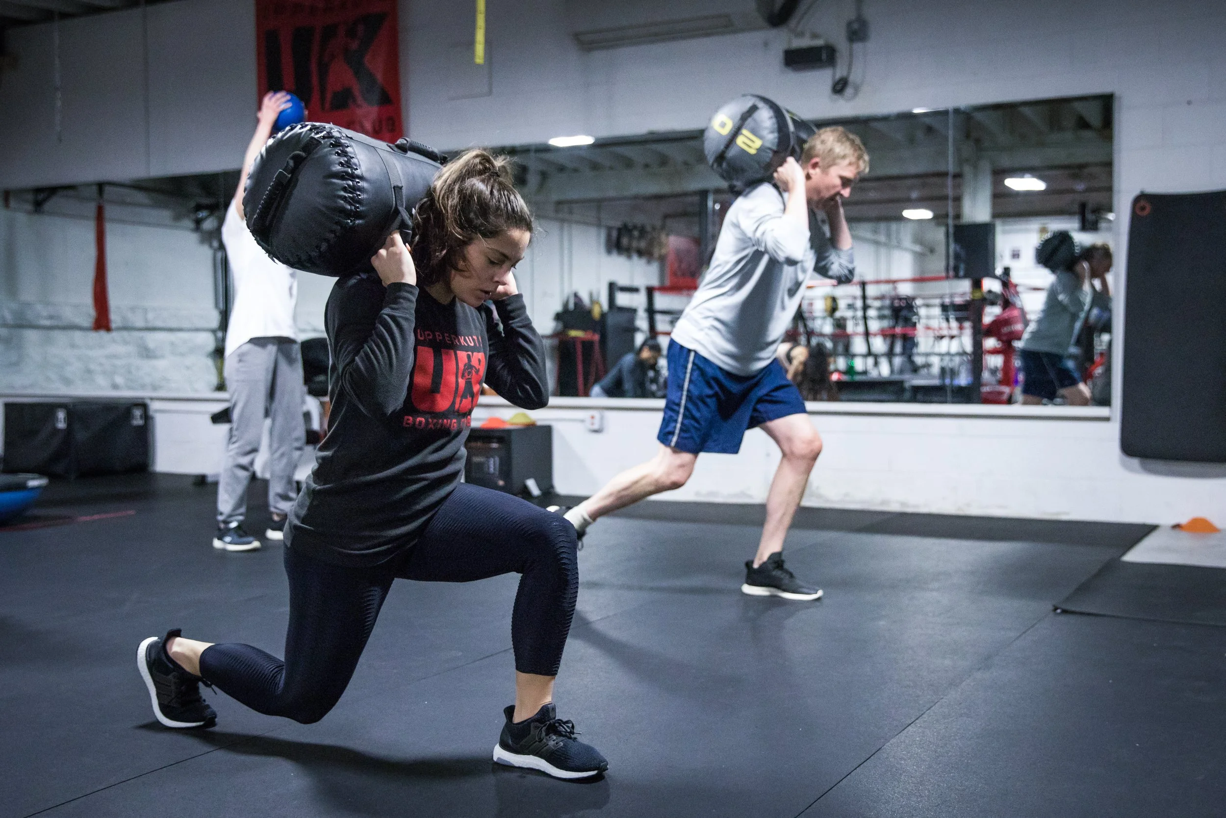 Two people working out in a gym, carrying weighted bags on their shoulders. The woman in the foreground is lunging forward, and the man in the background is stepping with a bag on his shoulders.