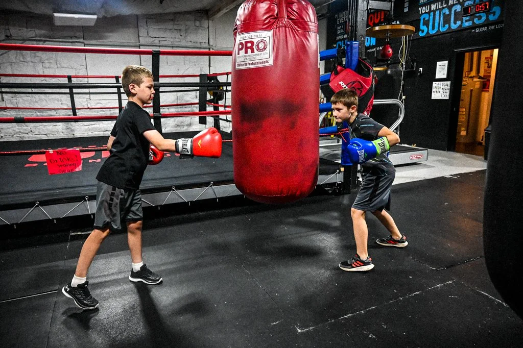 Two young boys practicing boxing with gloves on in a gym, hitting a large red punching bag.