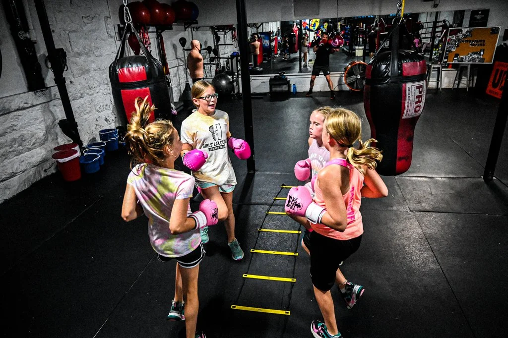Four young girls in athletic gear and pink boxing gloves participating in a boxing training session in a gym with punching bags and boxing equipment.