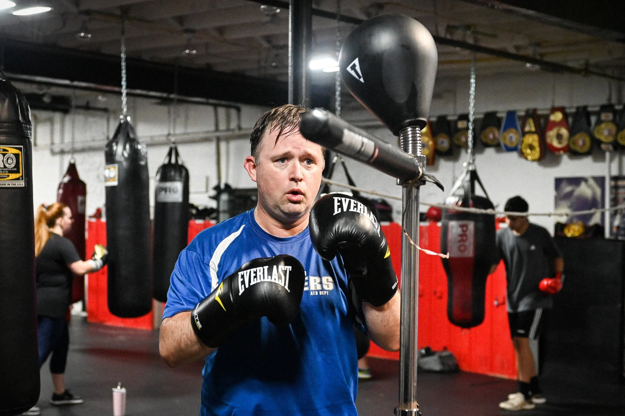 A man in blue sports attire and boxing gloves practicing punch on a speed bag in a boxing gym with other people in the background.