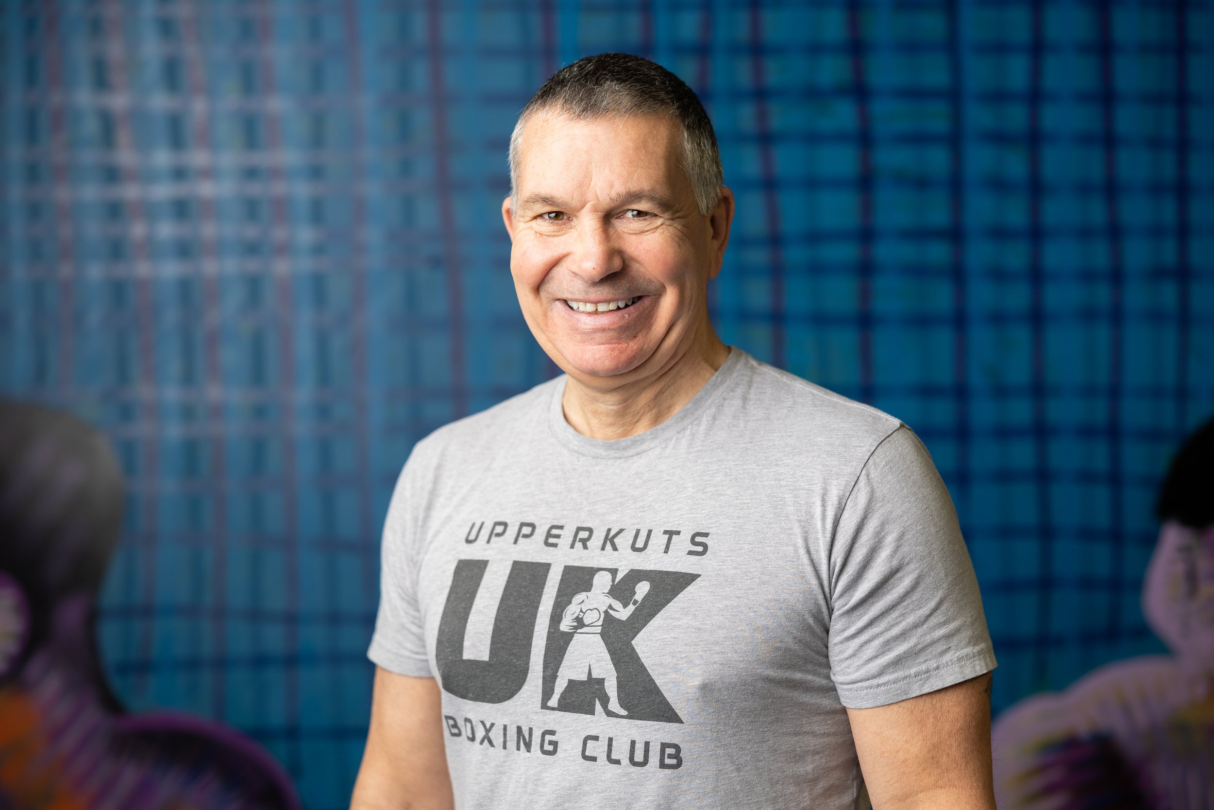 Smiling middle-aged man in a gray T-shirt with 'UpperKuts UK Boxing Club' logo, standing in front of a blue wall, with blurred figures in the background.