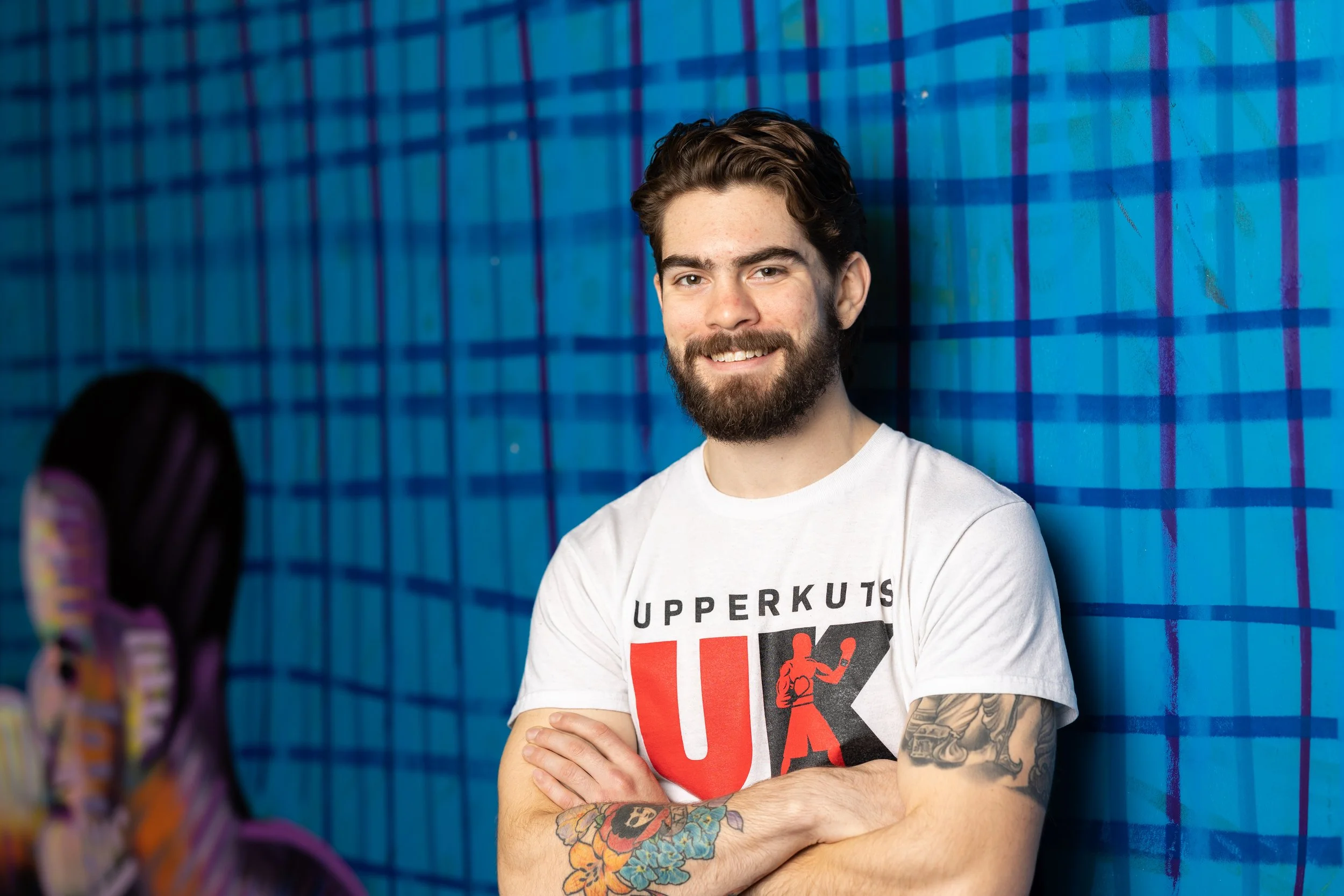 A young man with a beard and tattoos, smiling with arms crossed, standing against a blue plaid background, wearing a white T-shirt with 