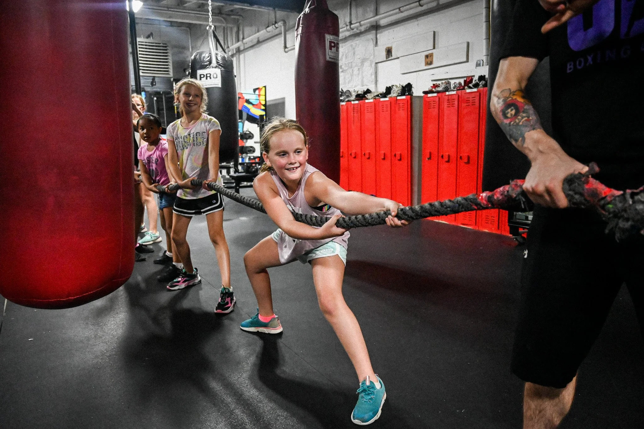 Children participating in a tug-of-war game with a trainer in a boxing gym. The girl in front is pulling the rope showing effort, with others lined up behind her. The gym has black flooring, red lockers, and punching bags.