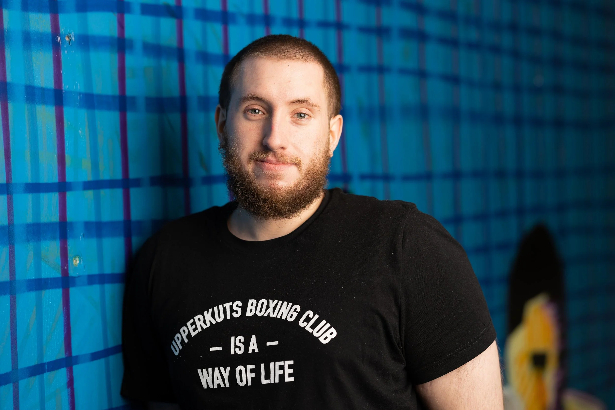 A young man with a beard and short hair standing against a colorful mural with a blue and purple checkered pattern, wearing a black T-shirt that reads 'UpperKuts Boxing Club is a way of life'.