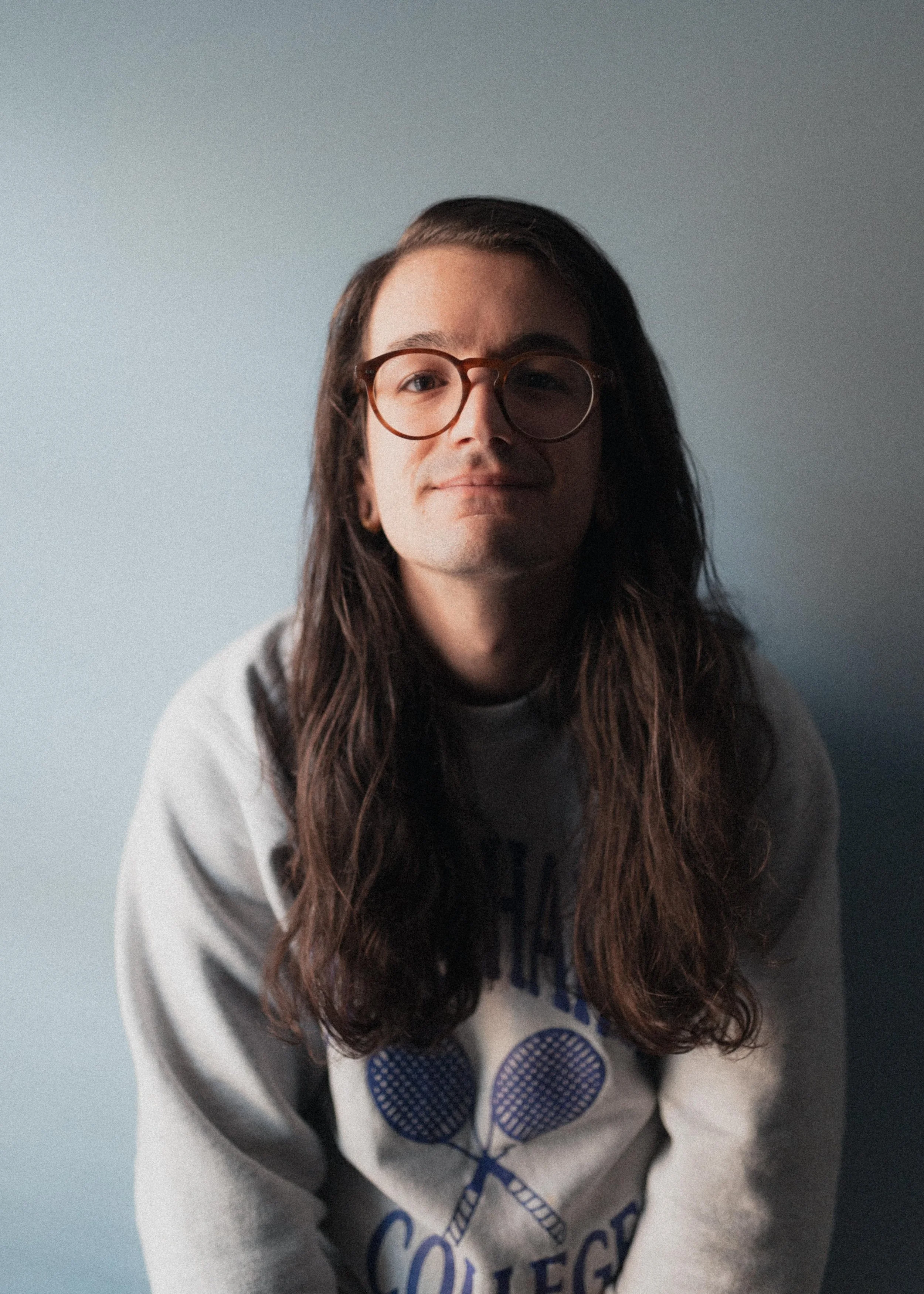 A young man with long brown hair, wearing glasses and a gray sweatshirt with tennis rackets and the word 'College' on it, sitting against a light blue wall.