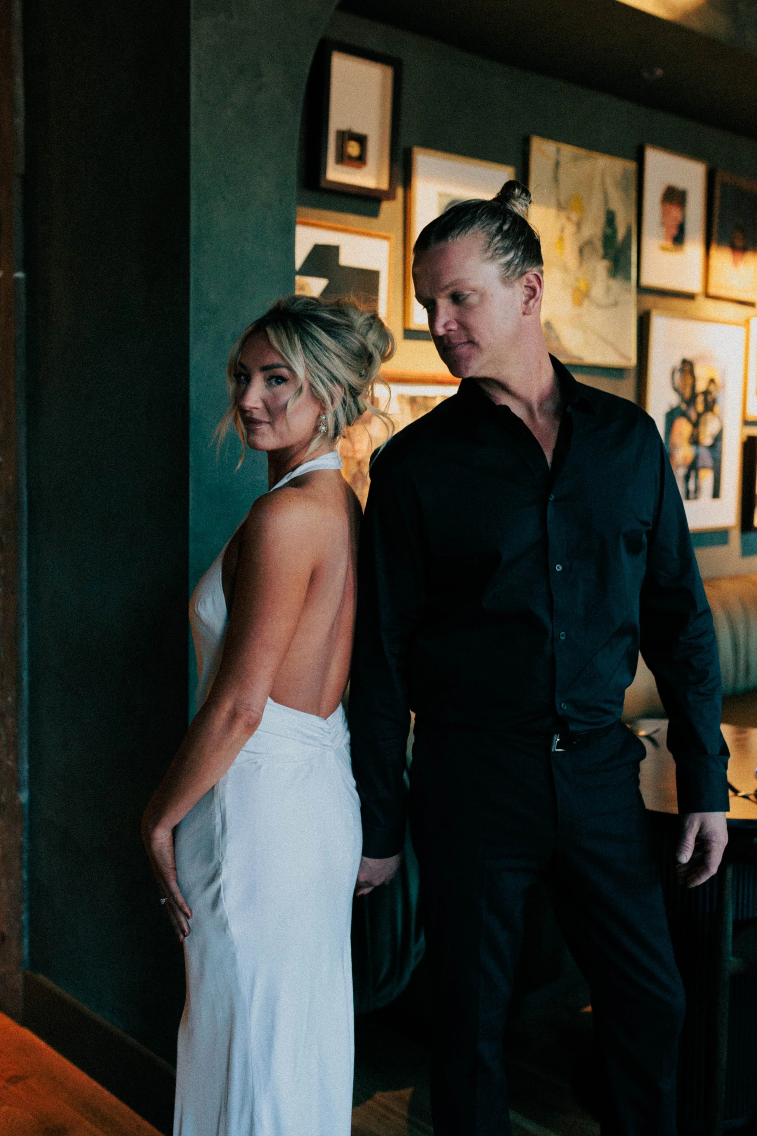 A man and a woman are standing back to back, holding hands, in an art gallery. The woman is wearing a white backless dress, and the man is dressed in black. They are indoors with framed artwork on the wall behind them.