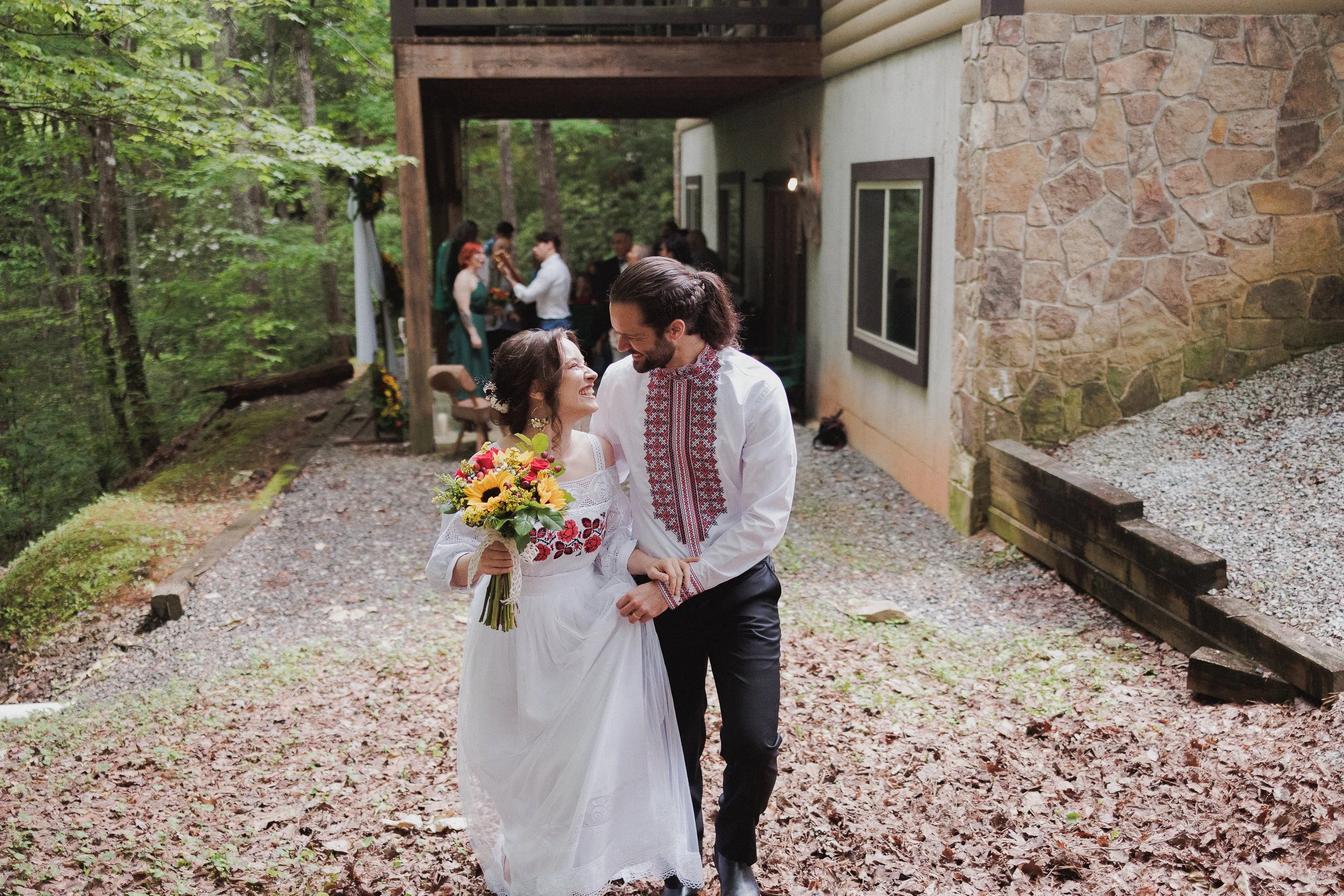 A newlywed couple dressed in traditional embroidered clothing walking outdoors, smiling and holding hands, with a bouquet of flowers, during a wedding celebration in a wooded area.