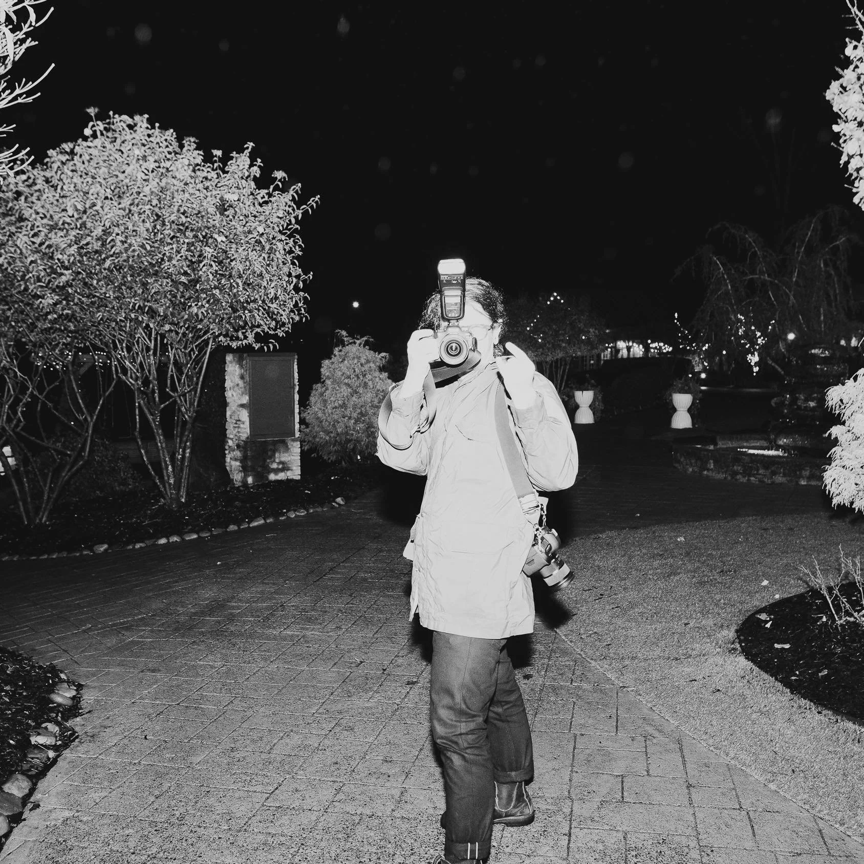 Black and white photo of a person standing on a paved path at night, holding a camera up to their face, with trees and decorative bushes in the background.